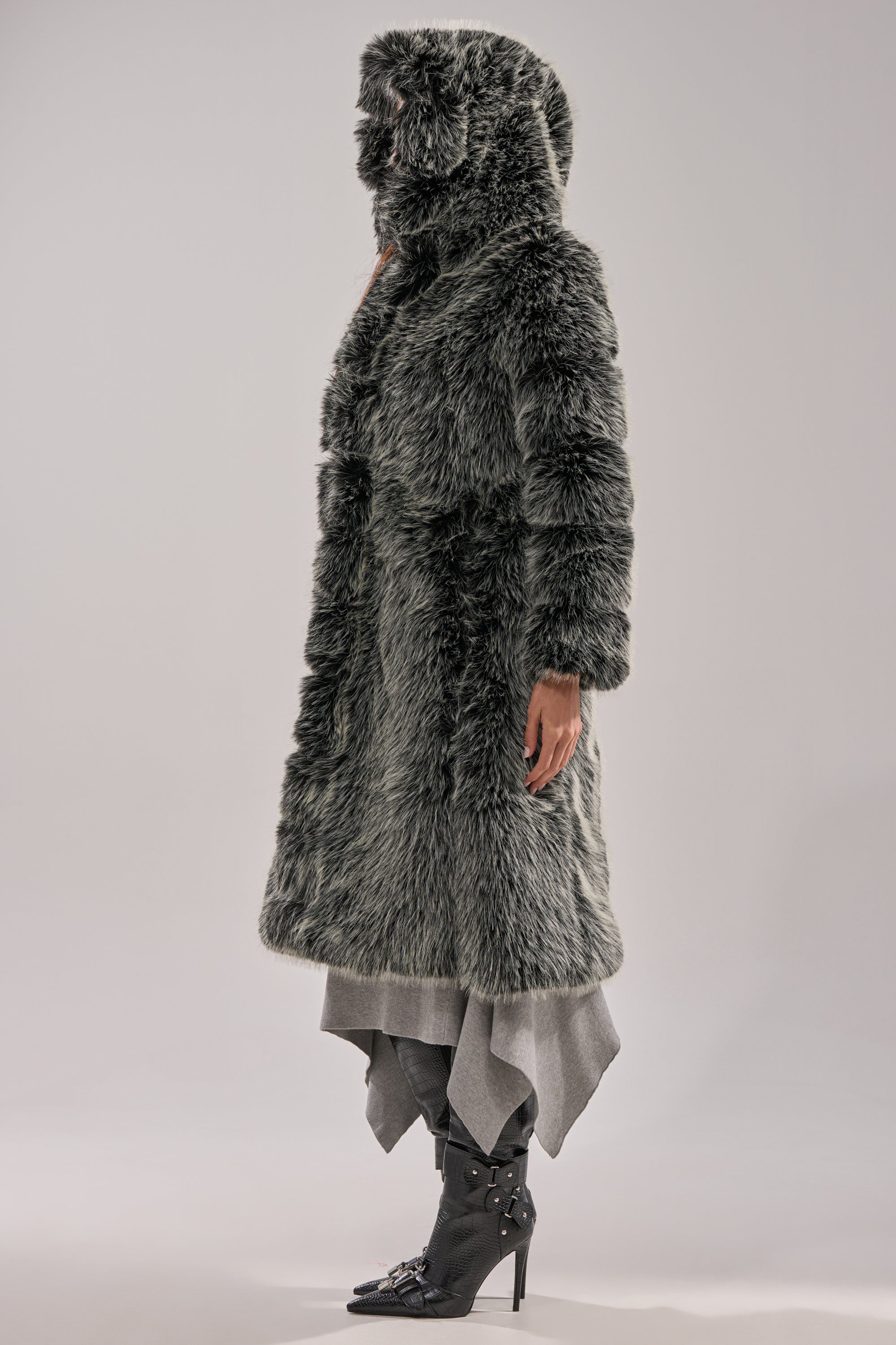 A person stands in profile against a plain light background, wearing the SNOW BUNNY HOODED FAUX FUR COAT in gray with a longline silhouette, layered skirt or pants, and black heeled boots.