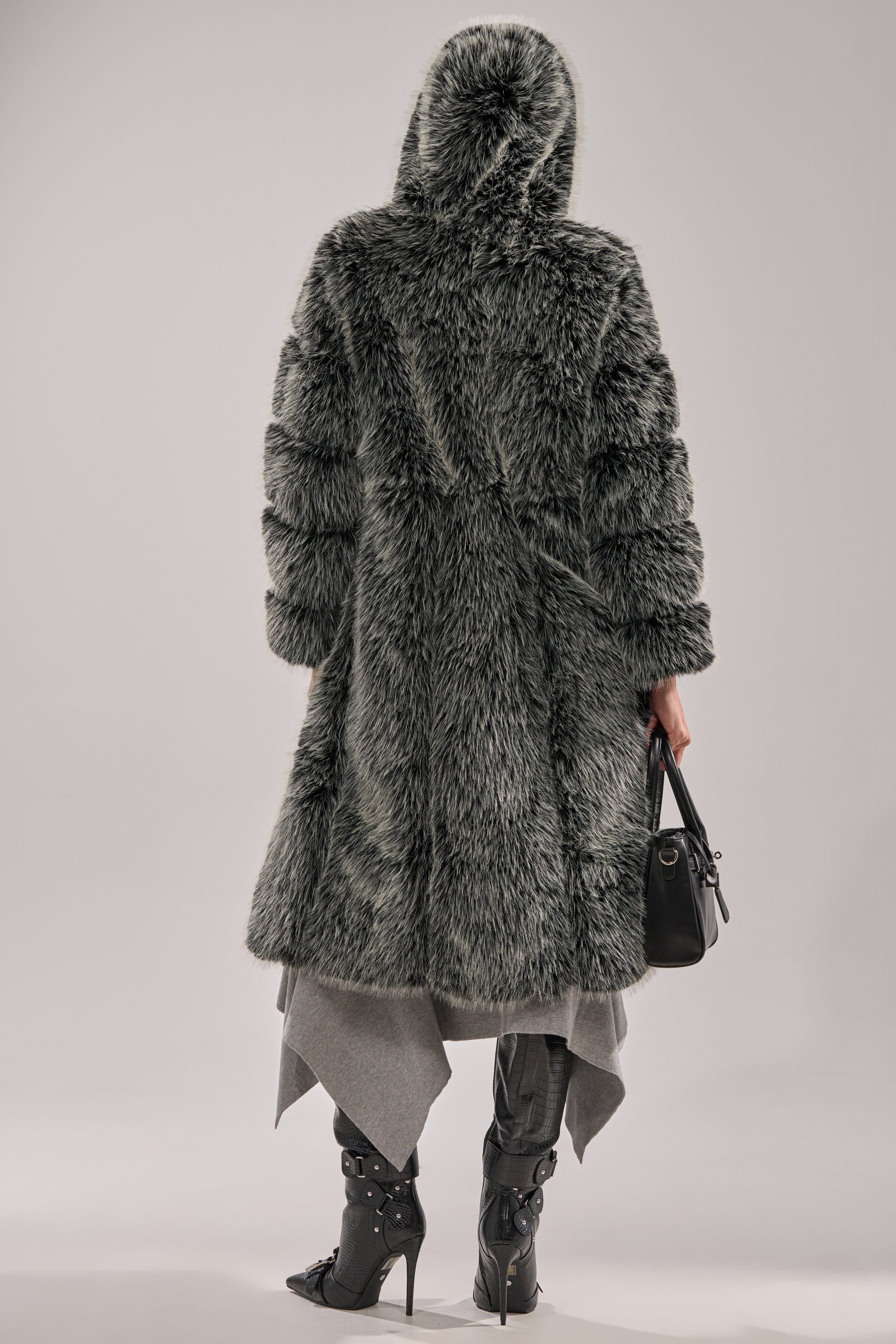 A person stands with their back turned, wearing the SNOW BUNNY HOODED FAUX FUR COAT in a longline style. They hold a black handbag, layer gray clothing, and wear black buckled heeled boots. The background is plain and light-colored.