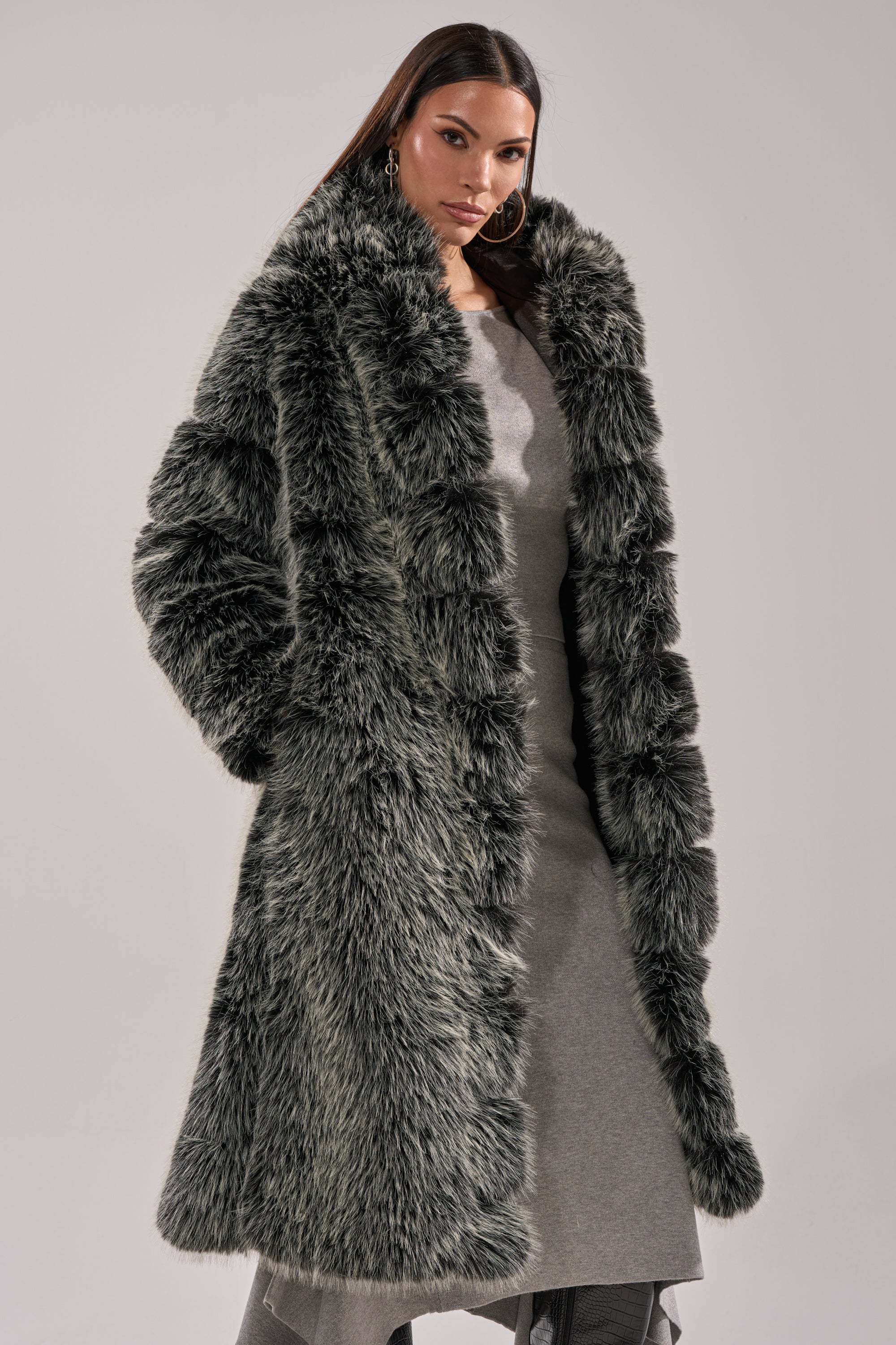 A woman wears the SNOW BUNNY HOODED FAUX FUR COAT over a gray dress, standing confidently with her hands in her pockets against a neutral background.