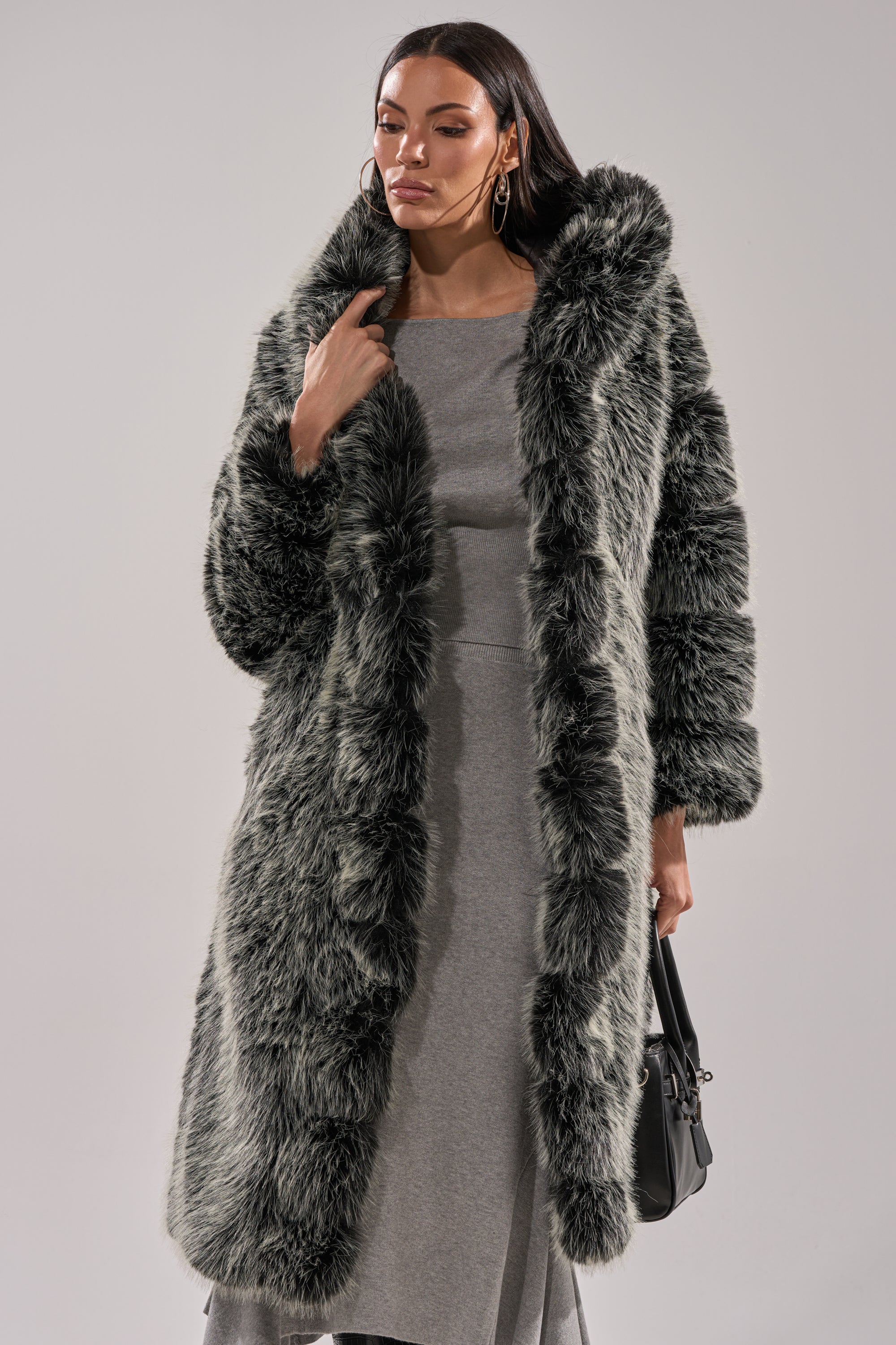 A woman stands against a plain background, confidently looking to the side while wearing the SNOW BUNNY HOODED FAUX FUR COAT over a light gray dress and holding a black handbag.