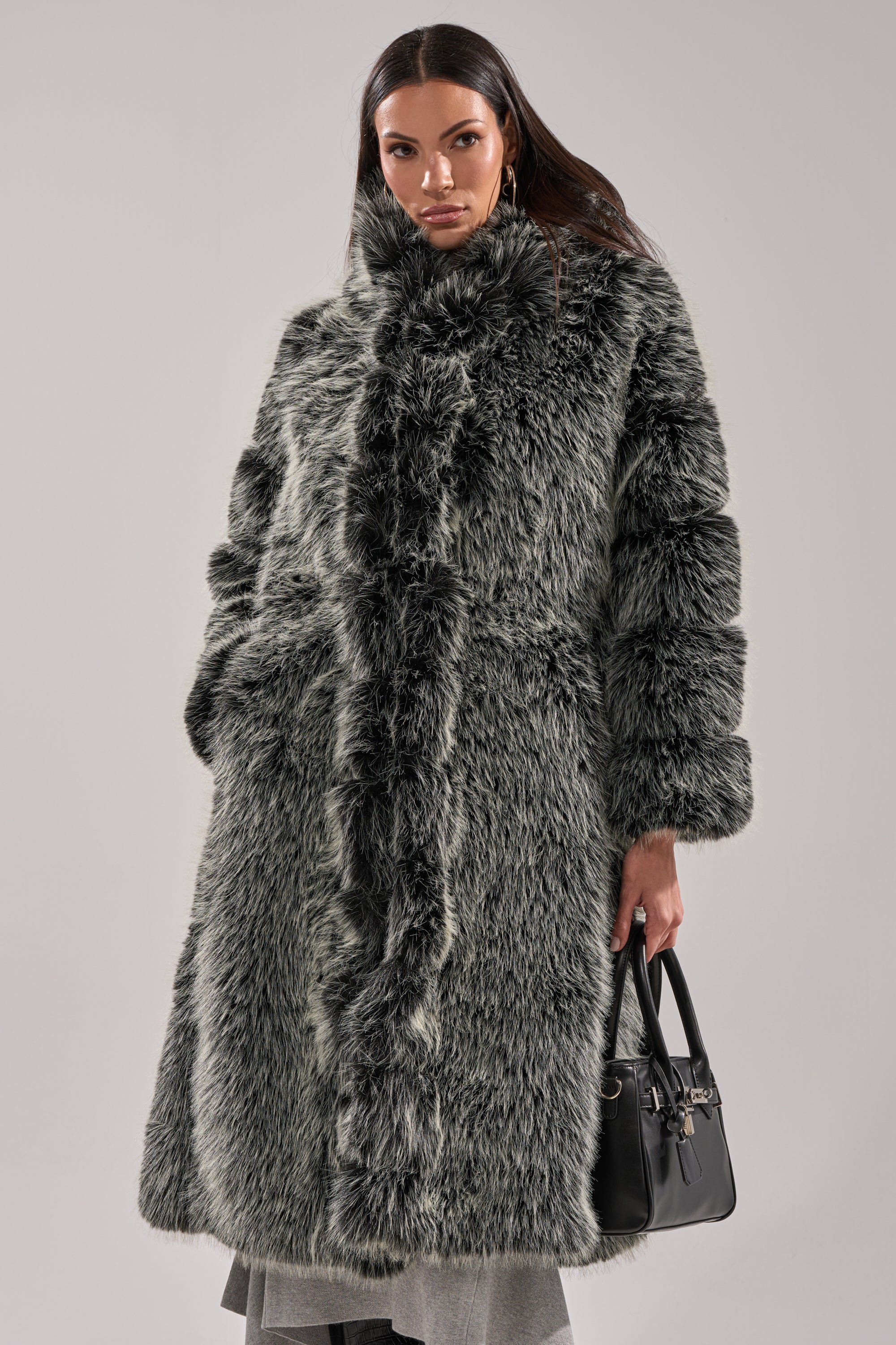 A woman poses confidently against a plain background, wearing the SNOW BUNNY HOODED FAUX FUR COAT and holding a black handbag in her left hand.