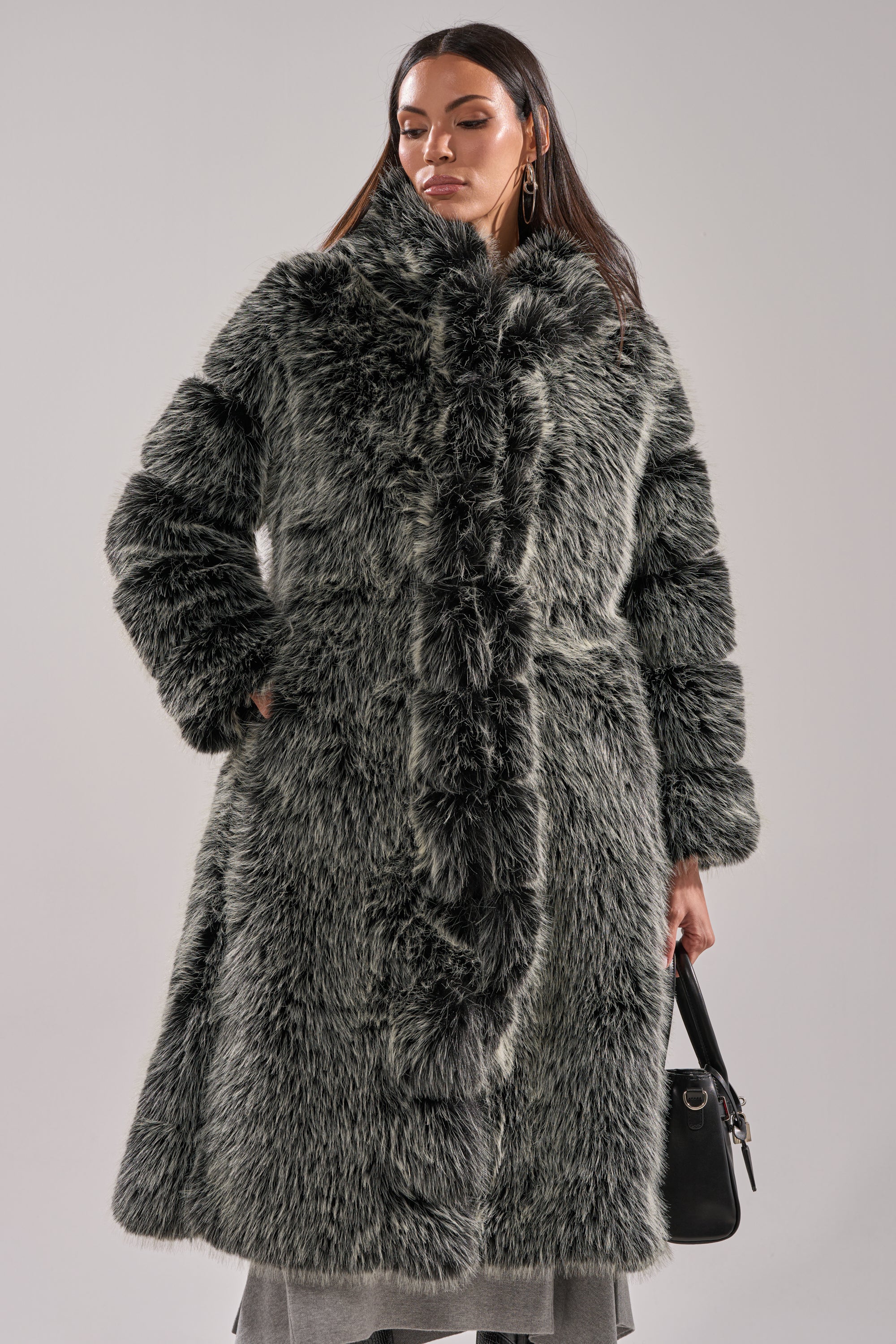 A woman poses against a light background, hands in pockets, wearing the SNOW BUNNY HOODED FAUX FUR COAT in gray. She accessorizes with gold hoop earrings, a black handbag, and has long dark hair.