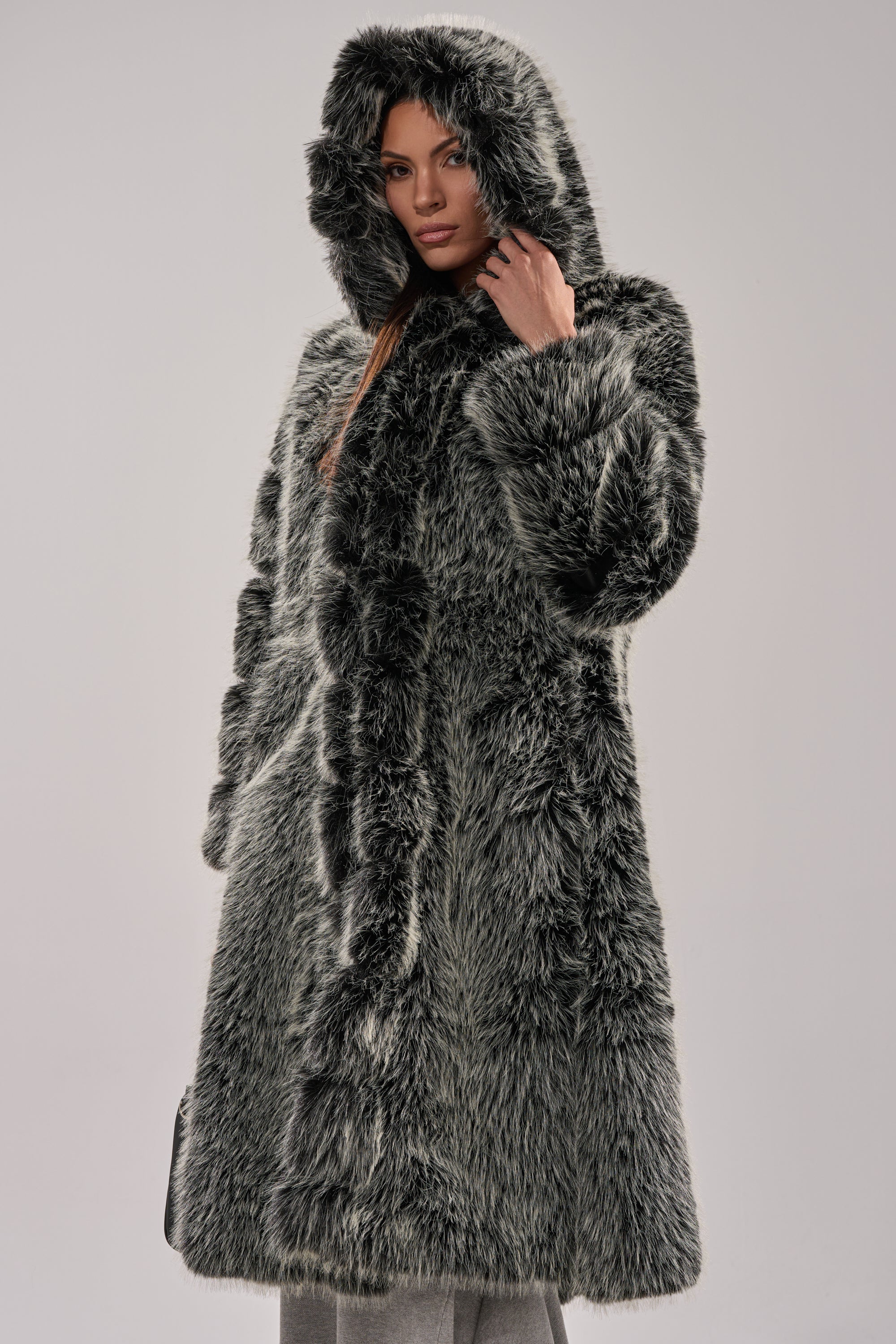 A woman models the SNOW BUNNY HOODED FAUX FUR COAT in thick gray, standing against a light background and facing forward as she gently touches the hood.