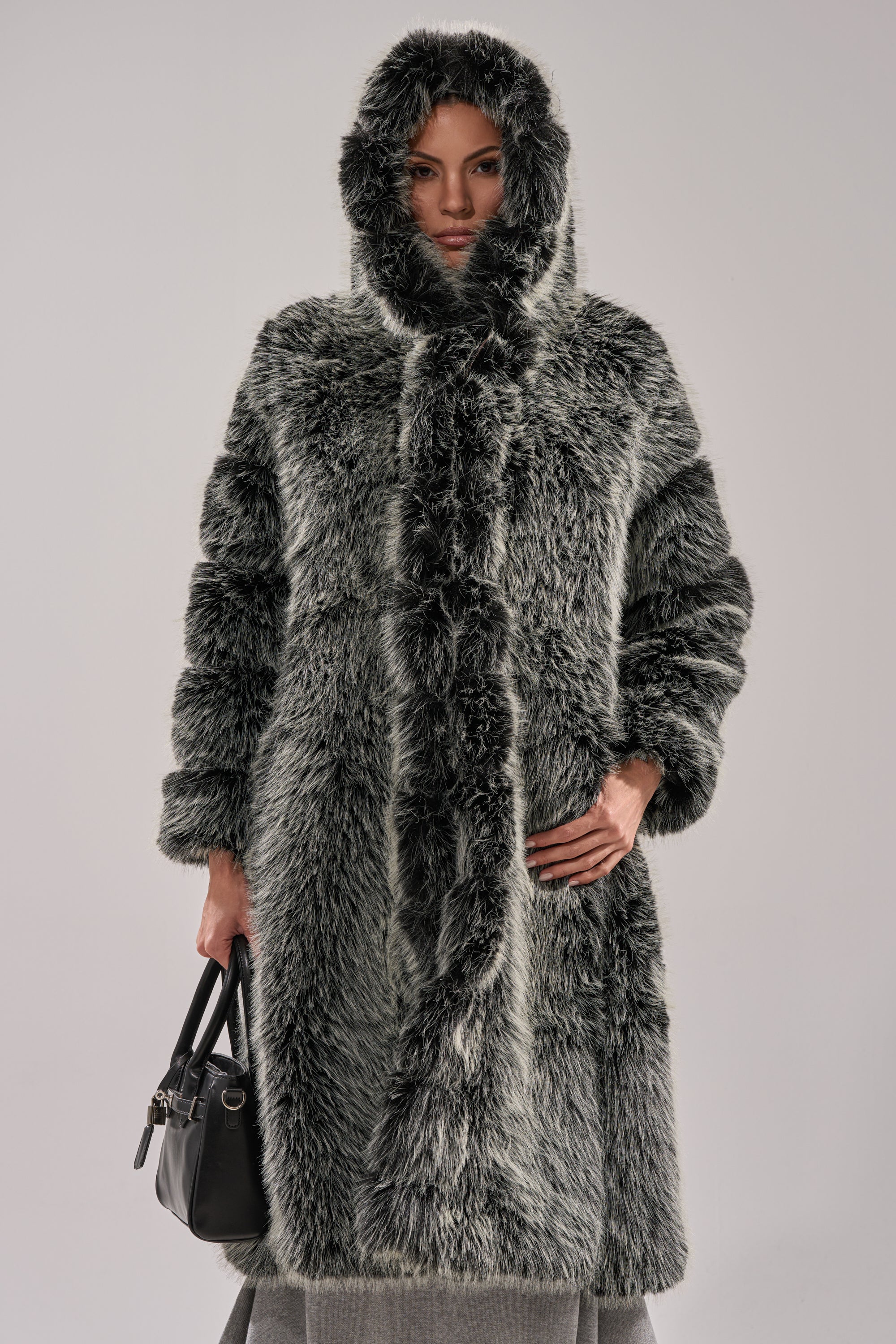 A person wearing the SNOW BUNNY HOODED FAUX FUR COAT with a longline silhouette and holding a black handbag stands against a plain light gray background.