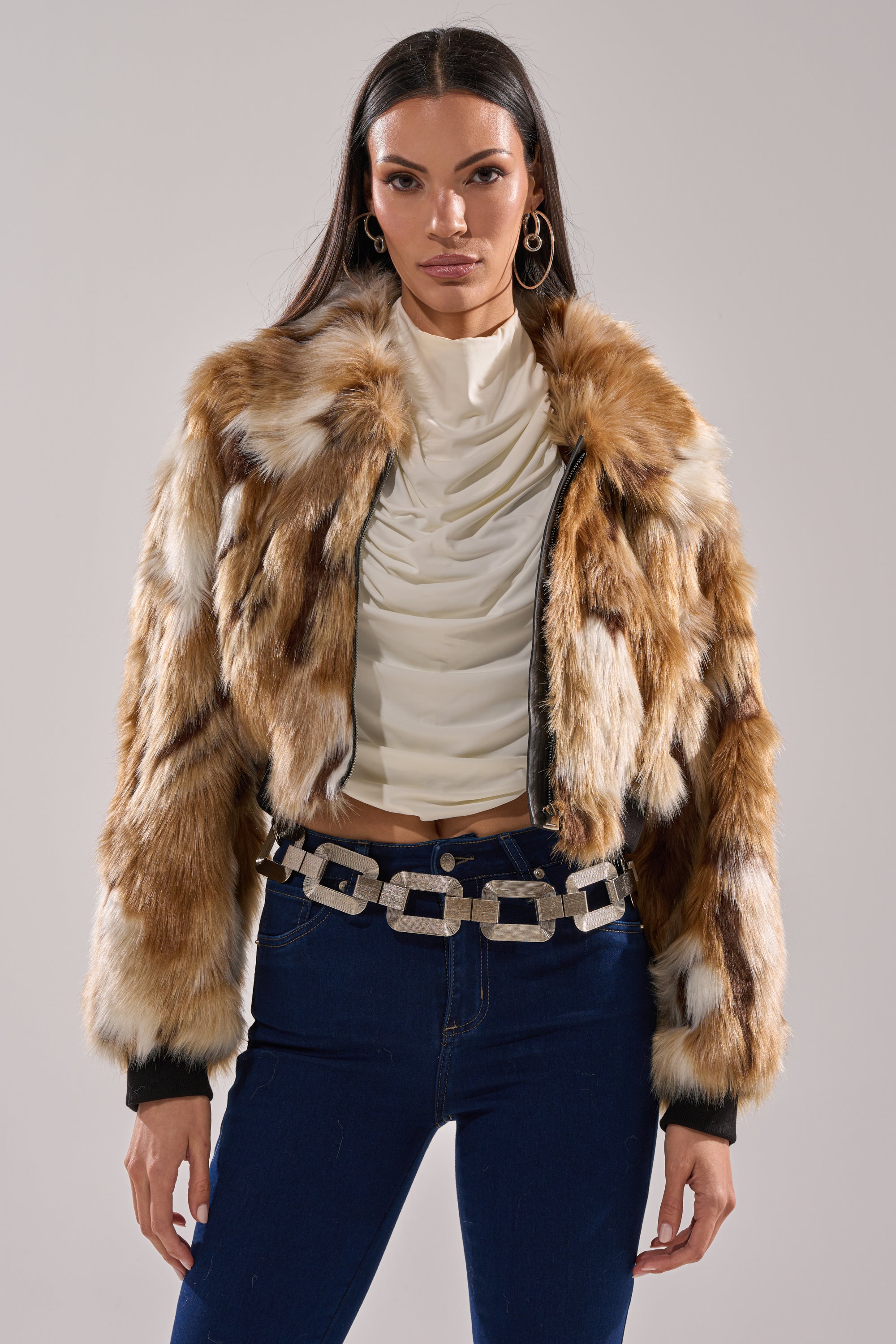 A woman stands facing forward, wearing the FENNEC FAUX FUR BOMBER in brown and cream, a white ruched top, blue jeans, a chunky silver chain belt, and large hoop earrings against a plain background.