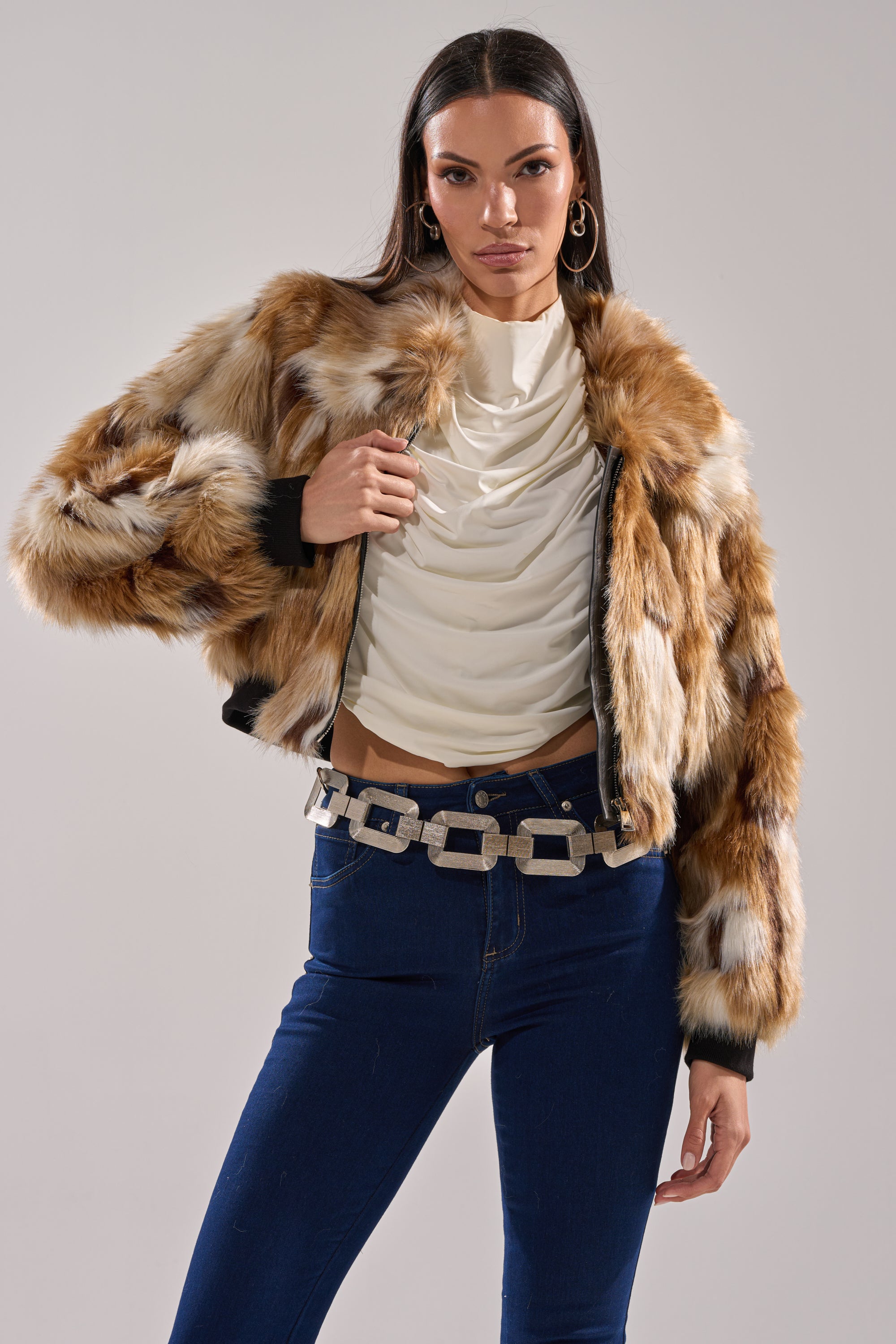 A woman poses confidently in the FENNEC FAUX FUR BOMBER, paired with a draped white top, dark blue jeans, and a chunky silver belt against a plain light background.