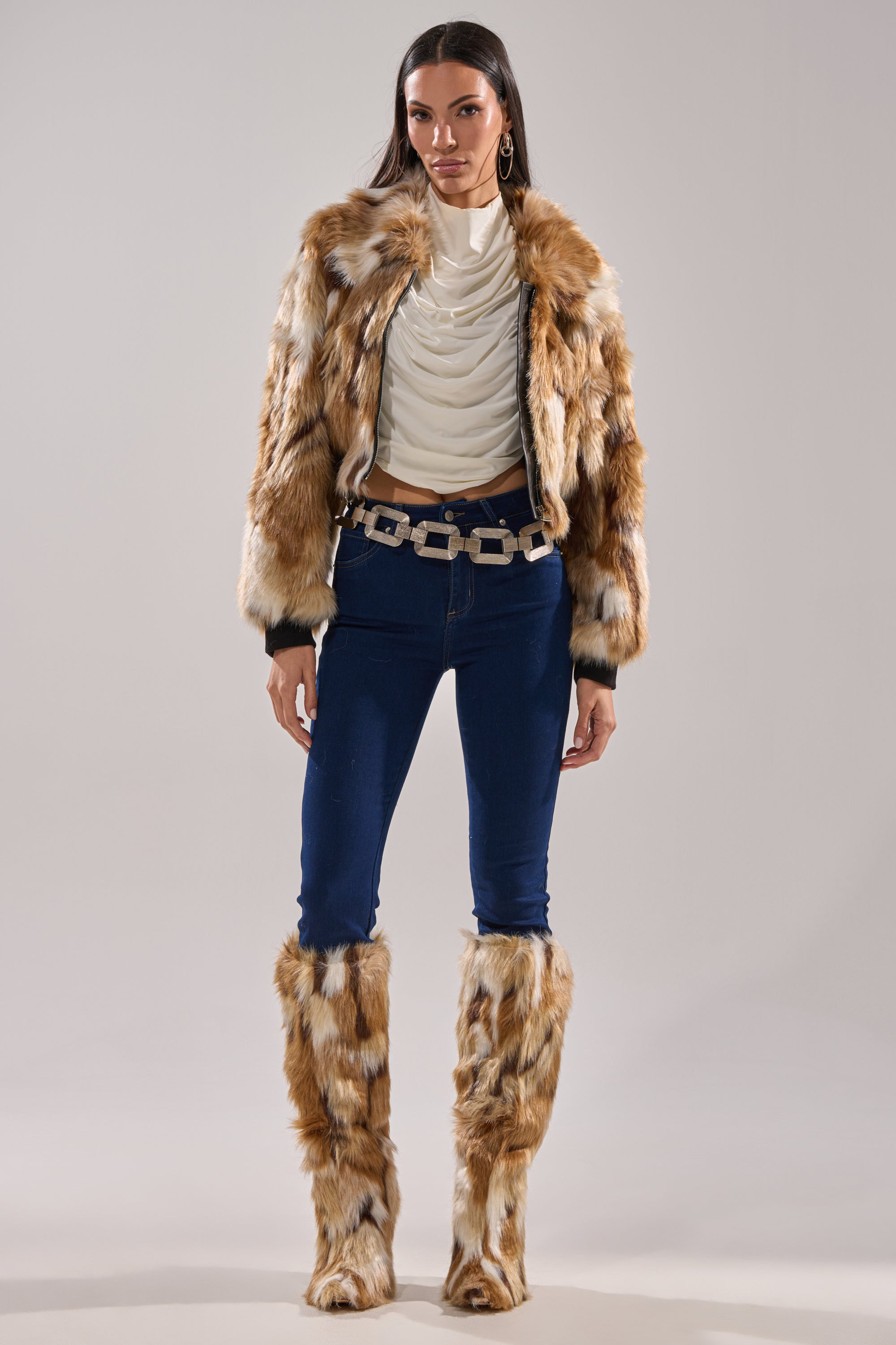 A woman stands facing forward against a plain background in the FENNEC FAUX FUR BOMBER with a cropped boxy fit, matching faux fur leg warmers, a white draped top, blue skinny jeans, hoop earrings, and a statement belt.