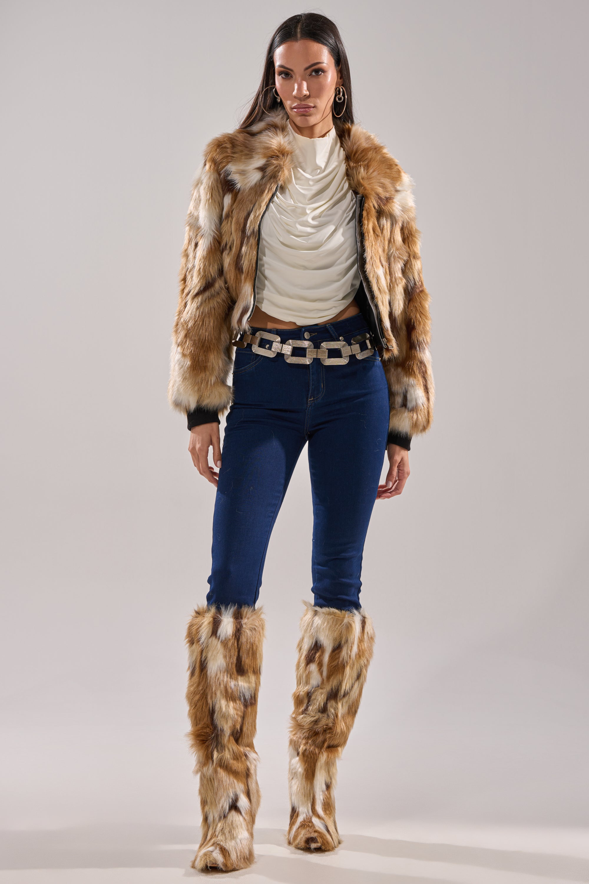 A woman wears the FENNEC FAUX FUR BOMBER with a cropped boxy fit, matching faux fur leg warmers, a white draped top, dark blue skinny jeans, a chunky chain belt, and hoop earrings against a plain background.