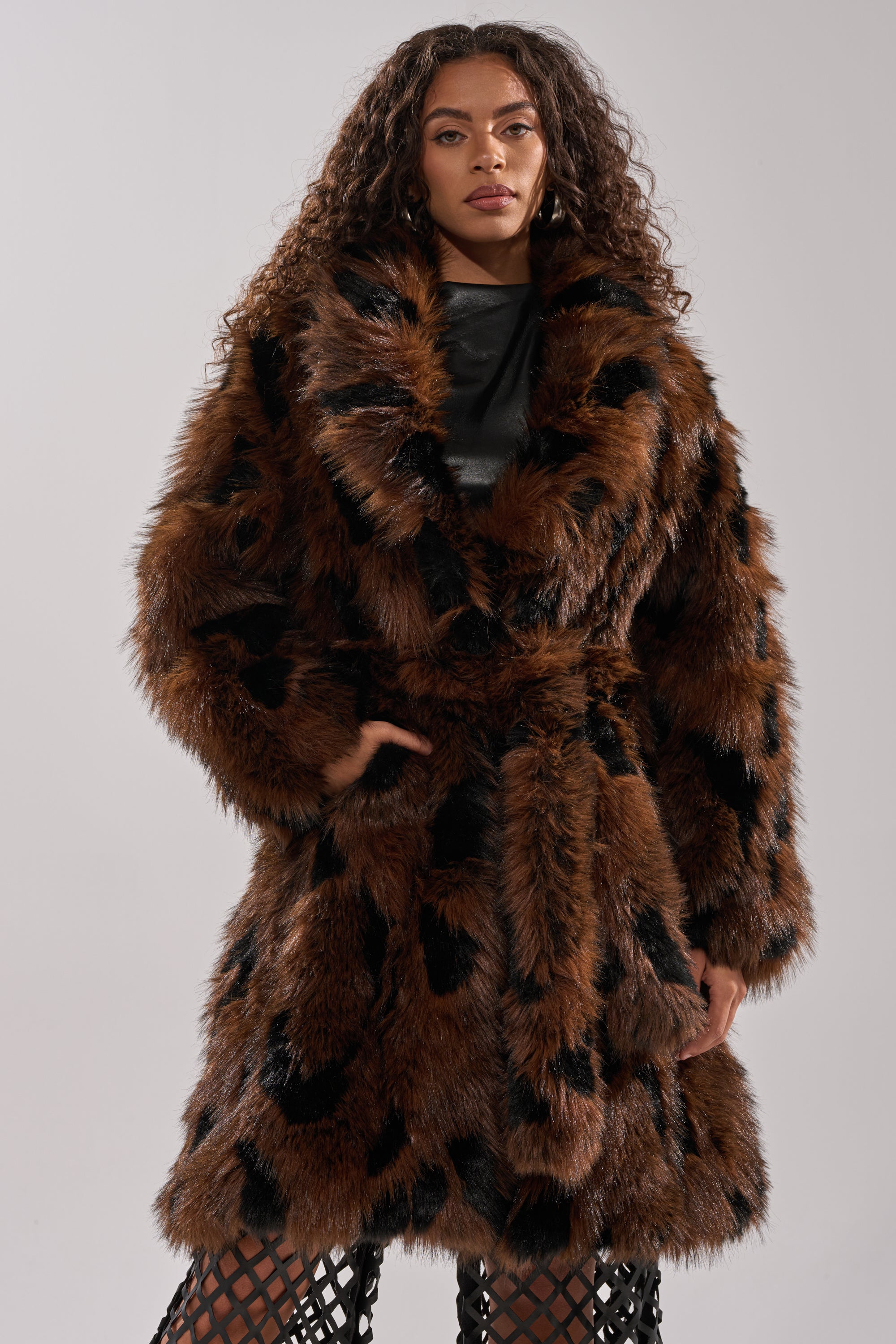 A woman with long curly hair exudes vintage glamour in the JUSTINE FAUX FUR COAT, featuring a brown and black design and belted waist, as she stands confidently with one hand in her pocket against a plain light background.