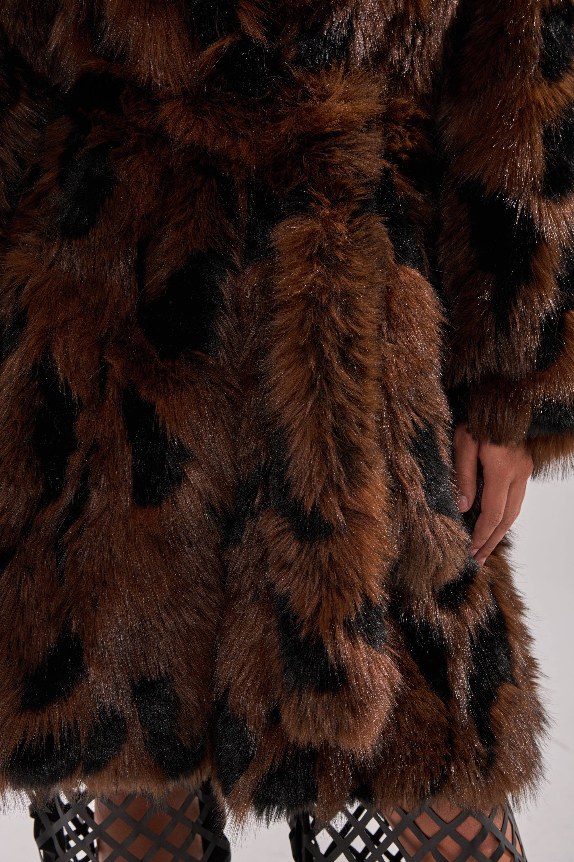 Close-up of a person wearing the JUSTINE FAUX FUR COAT, featuring brown faux fur with black spots for vintage glamour. Their hand rests by their side, paired with black fishnet stockings against a neutral background.