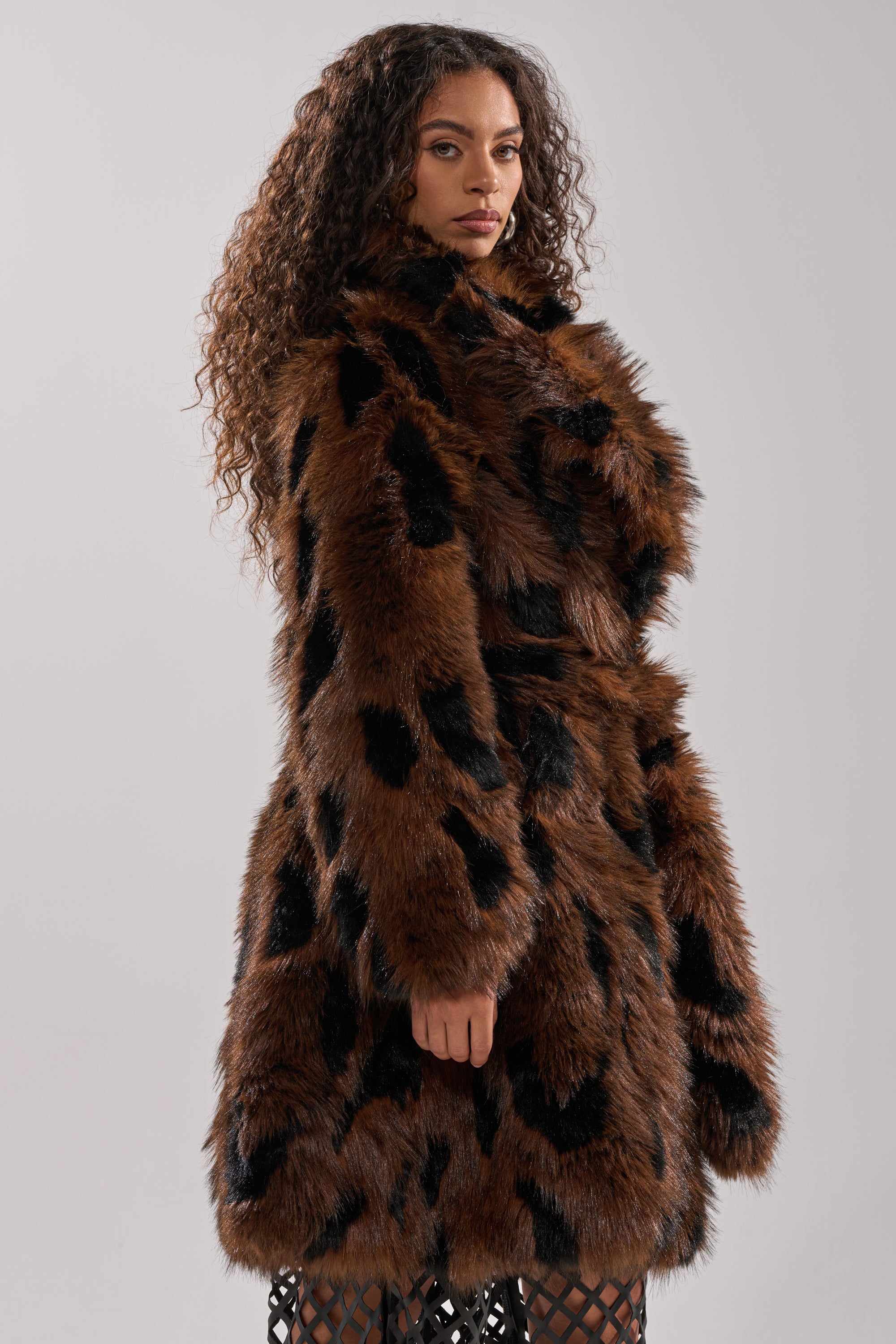 A woman with long, curly hair exudes vintage glamour in the JUSTINE FAUX FUR COAT, a brown longline style with black spots. She poses sideways, gazing at the camera against a plain light gray background.
