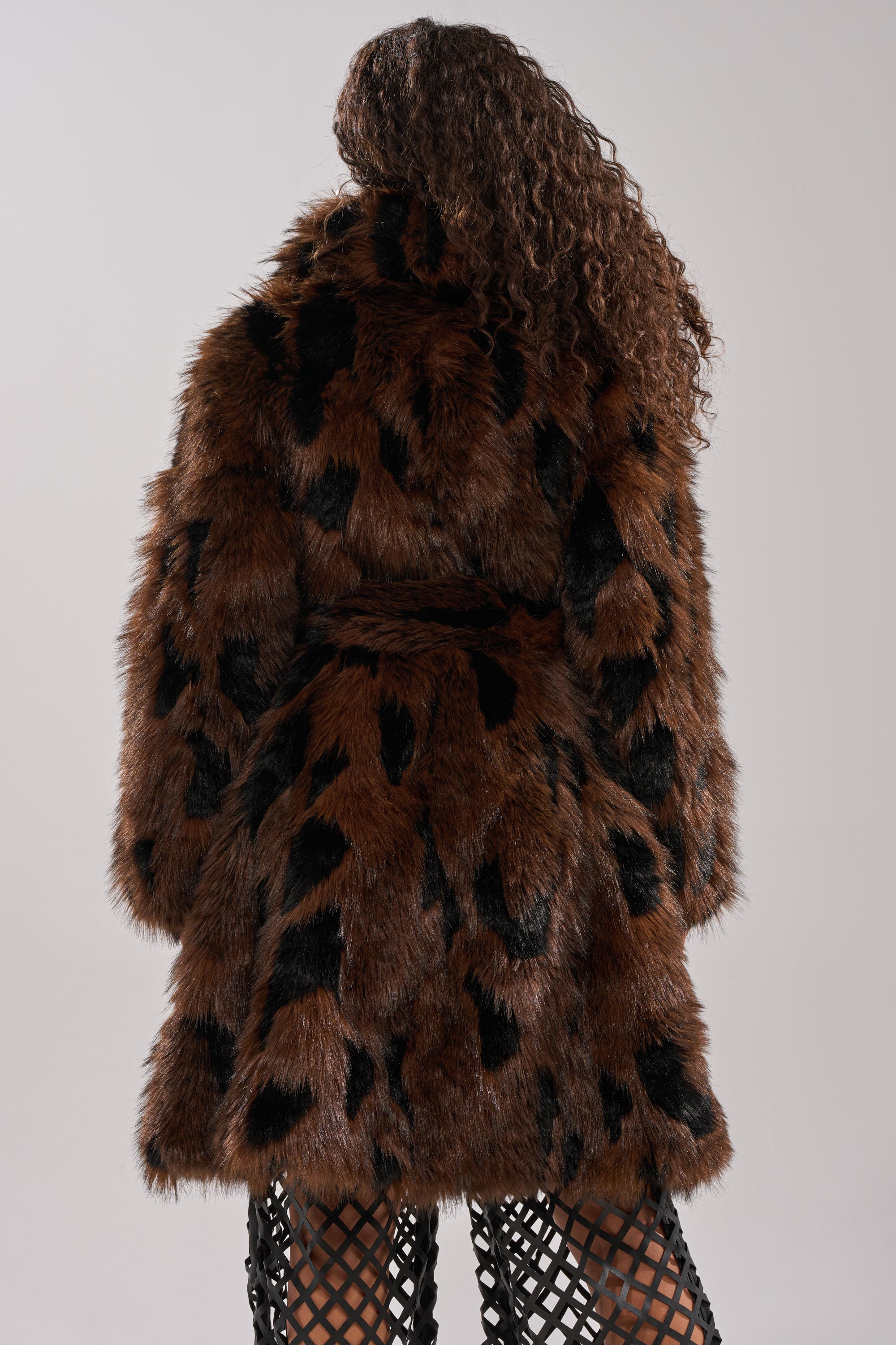 A person with curly hair models the JUSTINE FAUX FUR COAT—a brown longline piece with black spots and a belted waist—paired with black lattice-patterned leggings, shown from behind against a plain background.