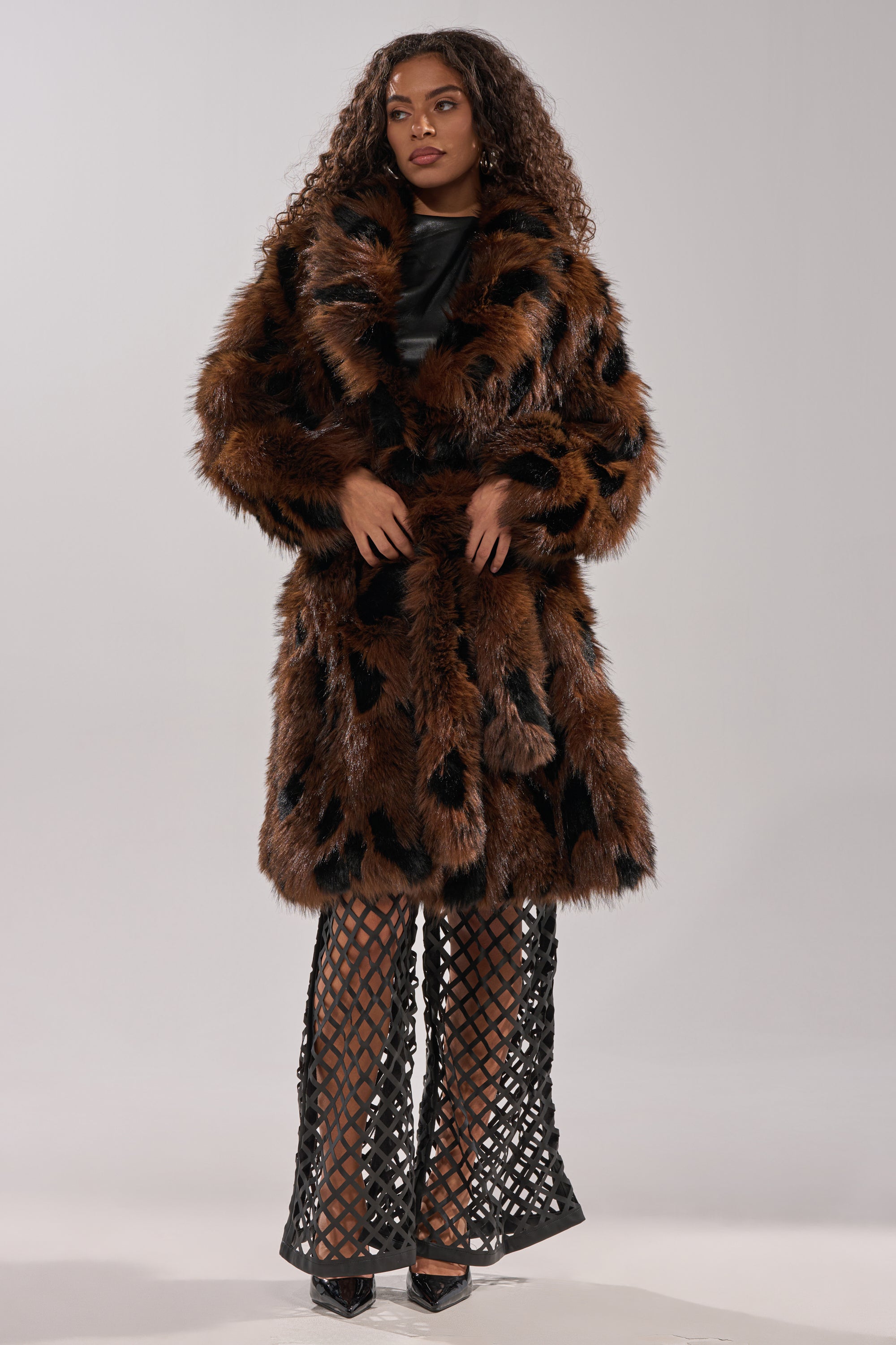 A woman with long curly hair exudes vintage glamour in the oversized brown and black JUSTINE FAUX FUR COAT over a black top, styled with high-waisted pants with crisscross cutouts and black pointed heels against a plain background.