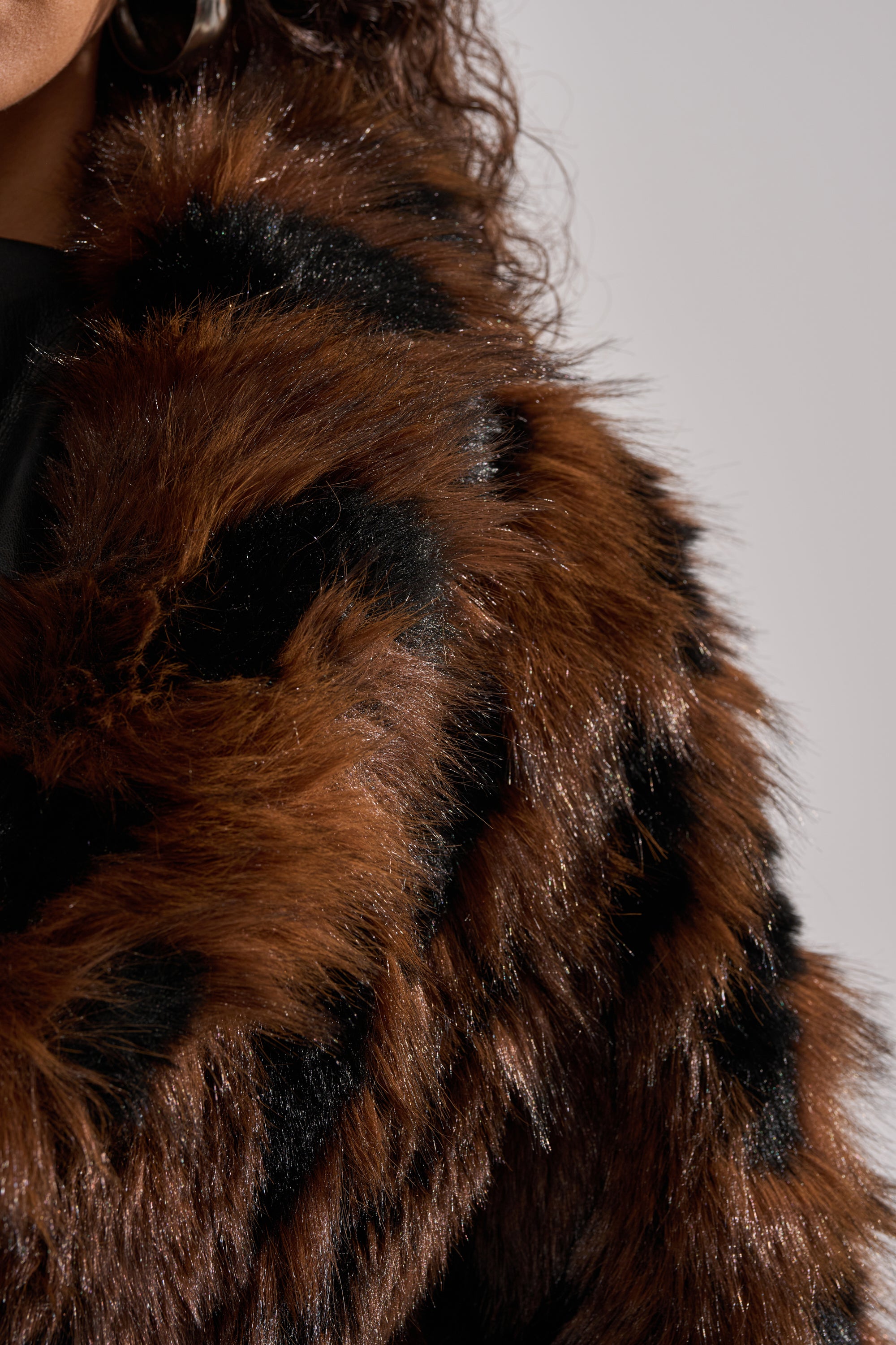 Close-up of a person in the JUSTINE FAUX FUR COAT, featuring brown and black faux fur with a shiny texture. Only part of their face, curly hair, and shoulder are visible against a neutral background, highlighting vintage glamour.