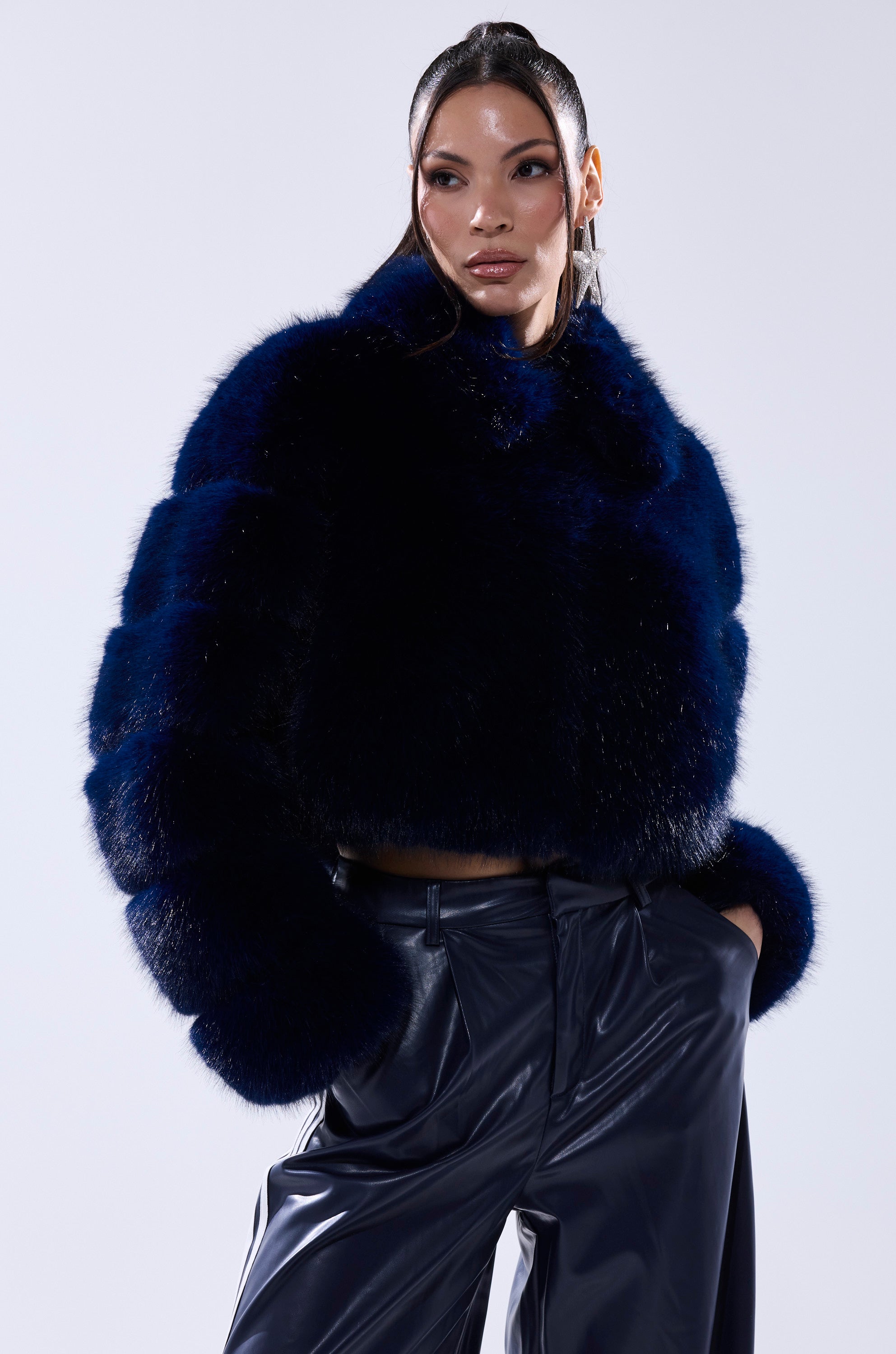 A woman exudes confidence in the KEIKO FAUX FUR COAT and sleek, high-waisted black pants, her pulled-back hair enhancing her bold, stylish look against a simple light background.