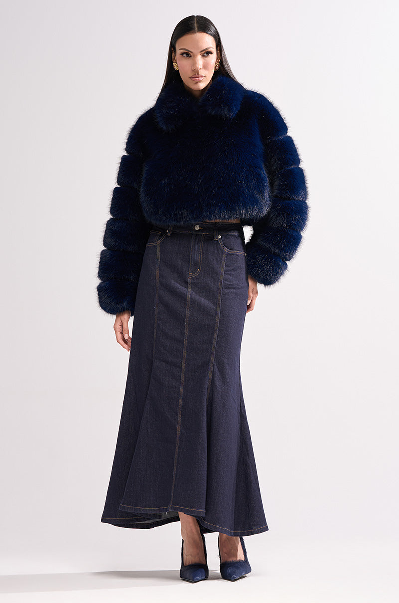 A woman poses against a plain backdrop wearing the KEIKO FAUX FUR COAT in dark blue, paired with a long flared denim skirt and pointed navy heels. She has sleek, straight hair and a neutral expression.