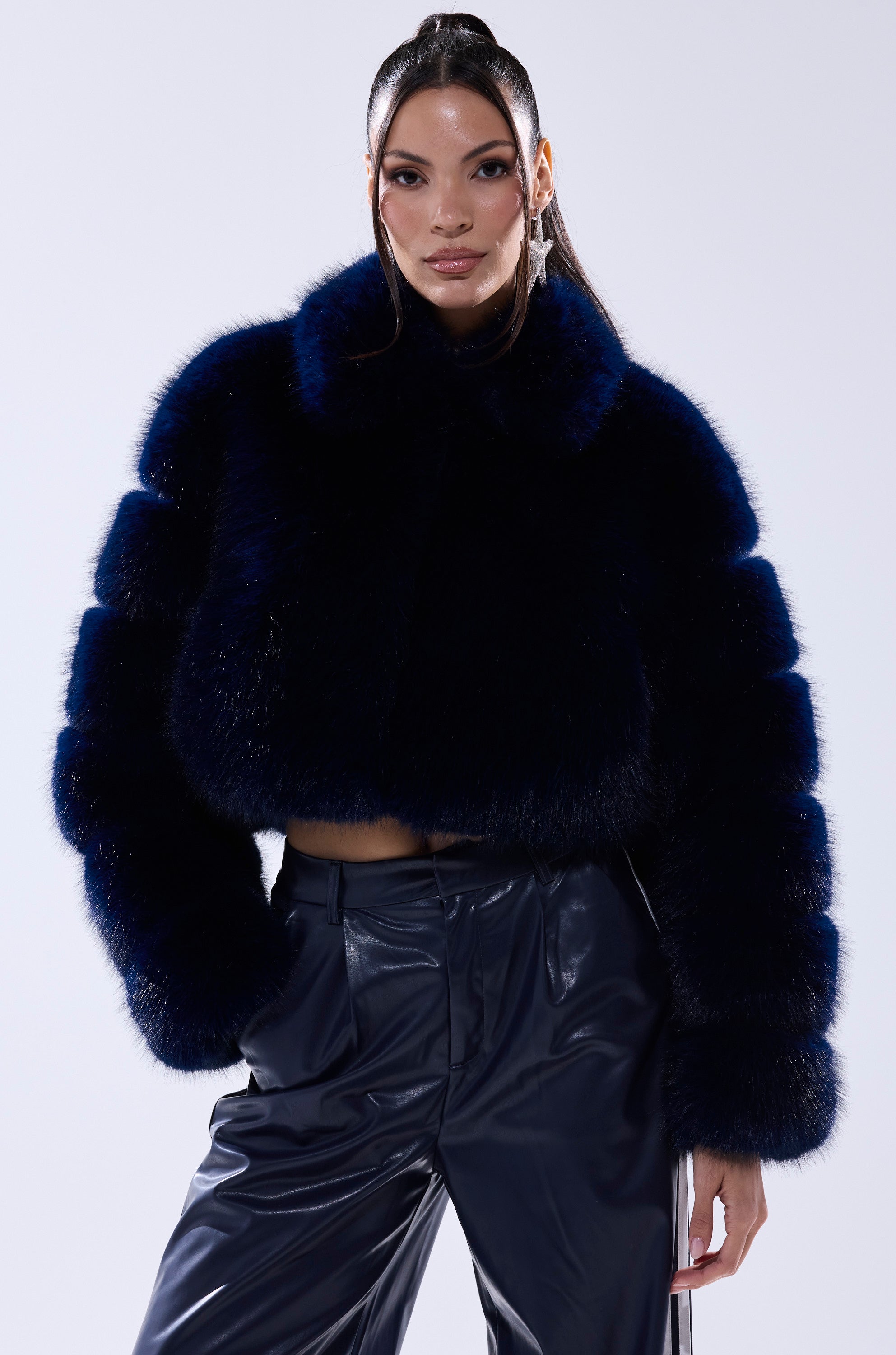 A woman poses confidently in the KEIKO FAUX FUR COAT, a striking dark blue cropped jacket, paired with shiny dark pants and her hand in her pocket, set against a plain light background.