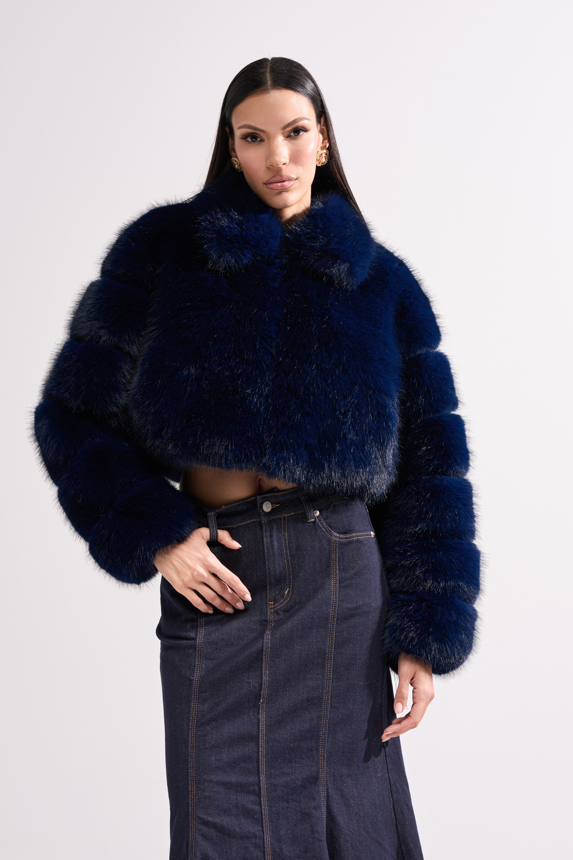 A woman with long dark hair wears the KEIKO FAUX FUR COAT in plush navy blue and a high-waisted dark denim skirt, standing against a plain light background.