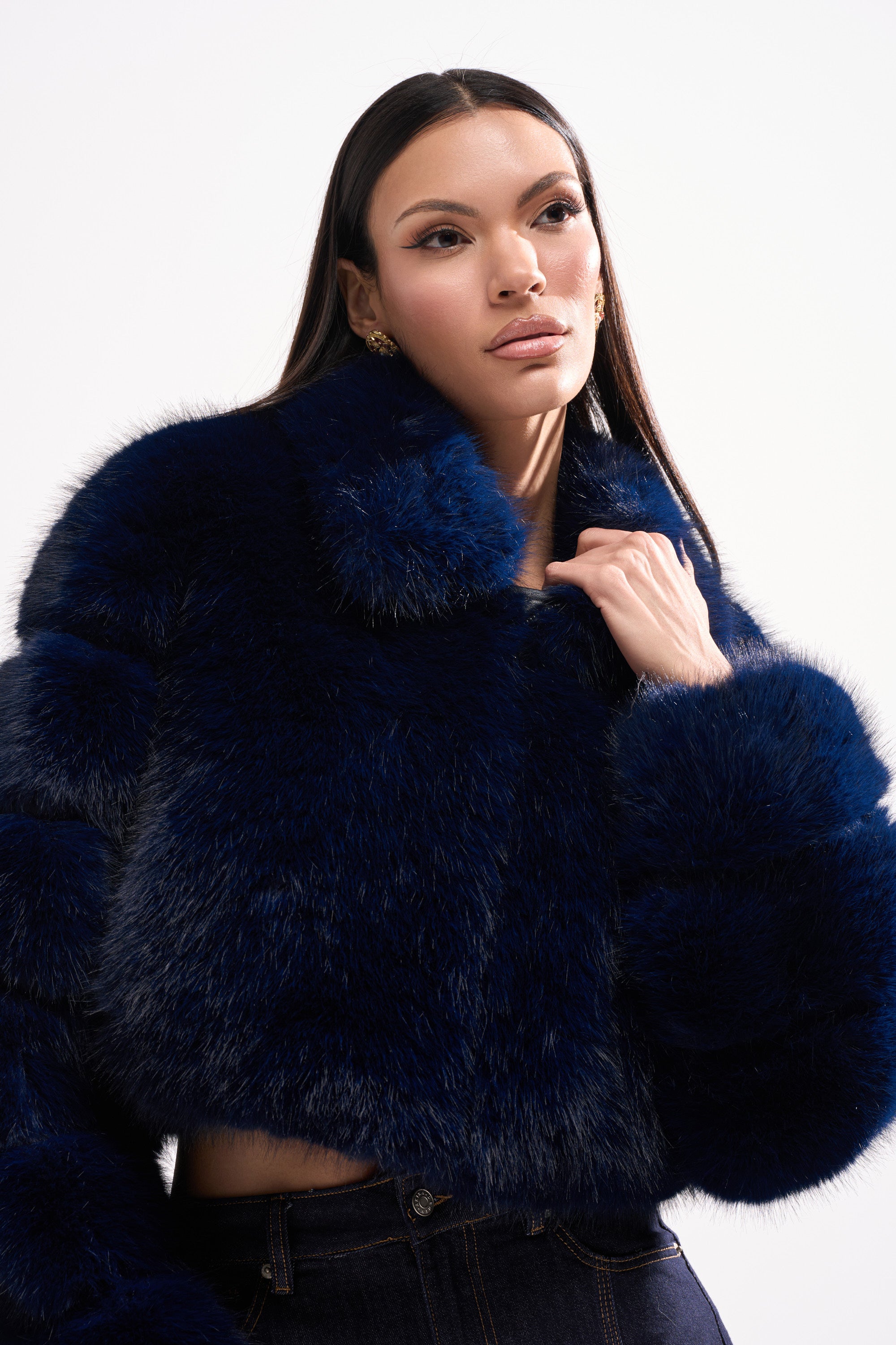 A woman with long straight hair models the KEIKO FAUX FUR COAT in deep blue with dark jeans, posing confidently against a white background.