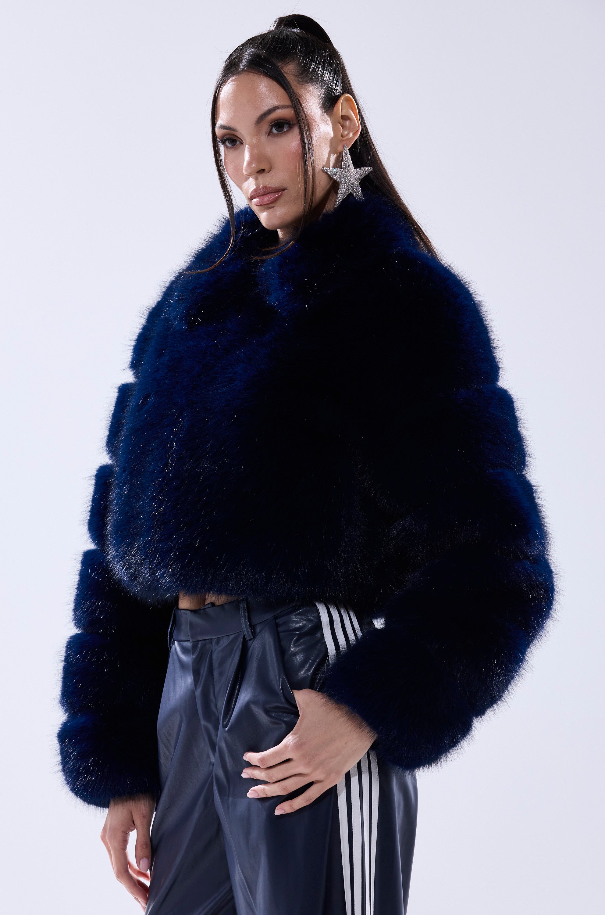 A woman wearing the KEIKO FAUX FUR COAT in dark blue, navy track pants with white stripes, and a large star-shaped earring poses against a plain light background.