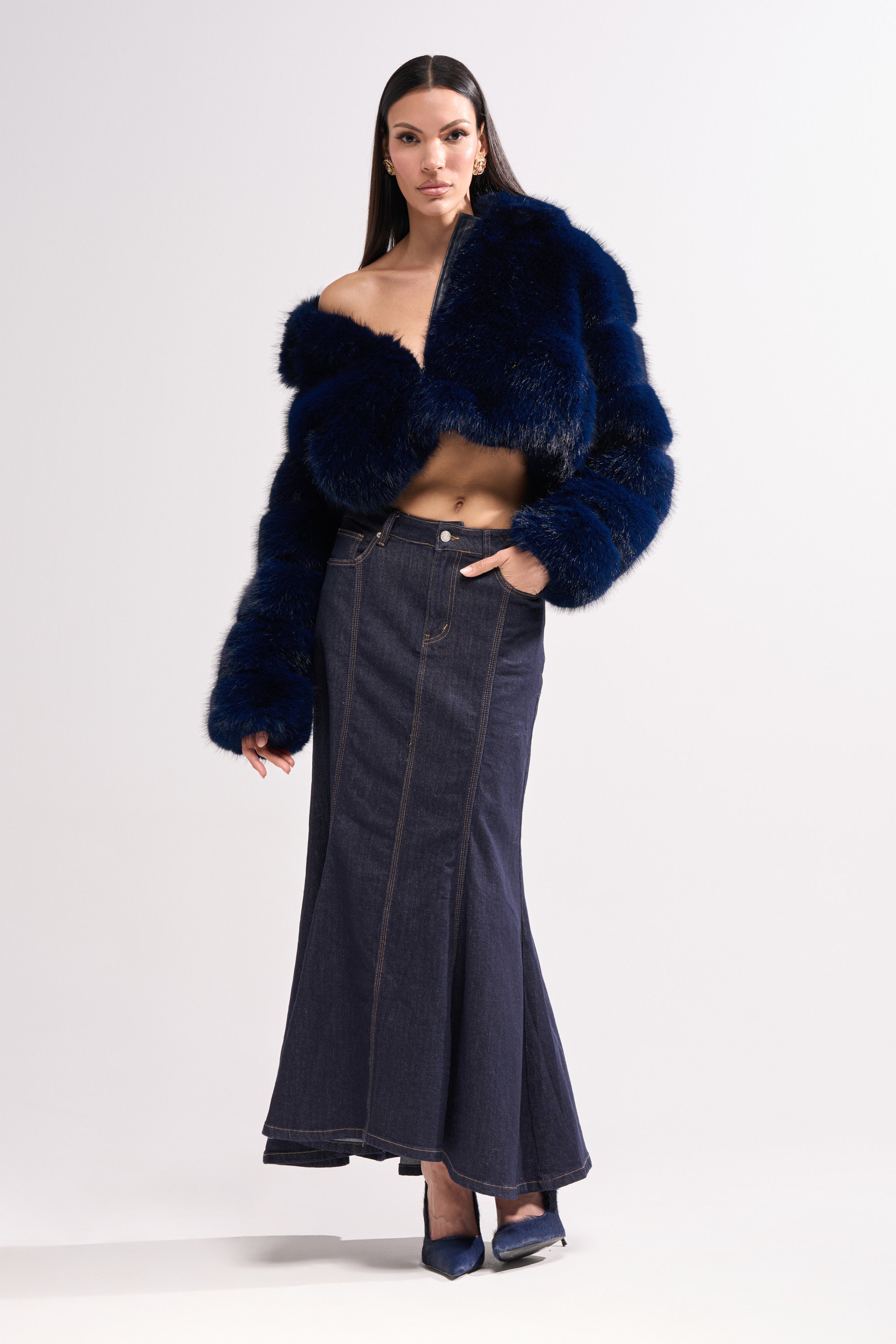 A woman models the KEIKO FAUX FUR COAT off the shoulder, styled with a high-waisted denim skirt and pointed-toe heels, set against a plain white background.