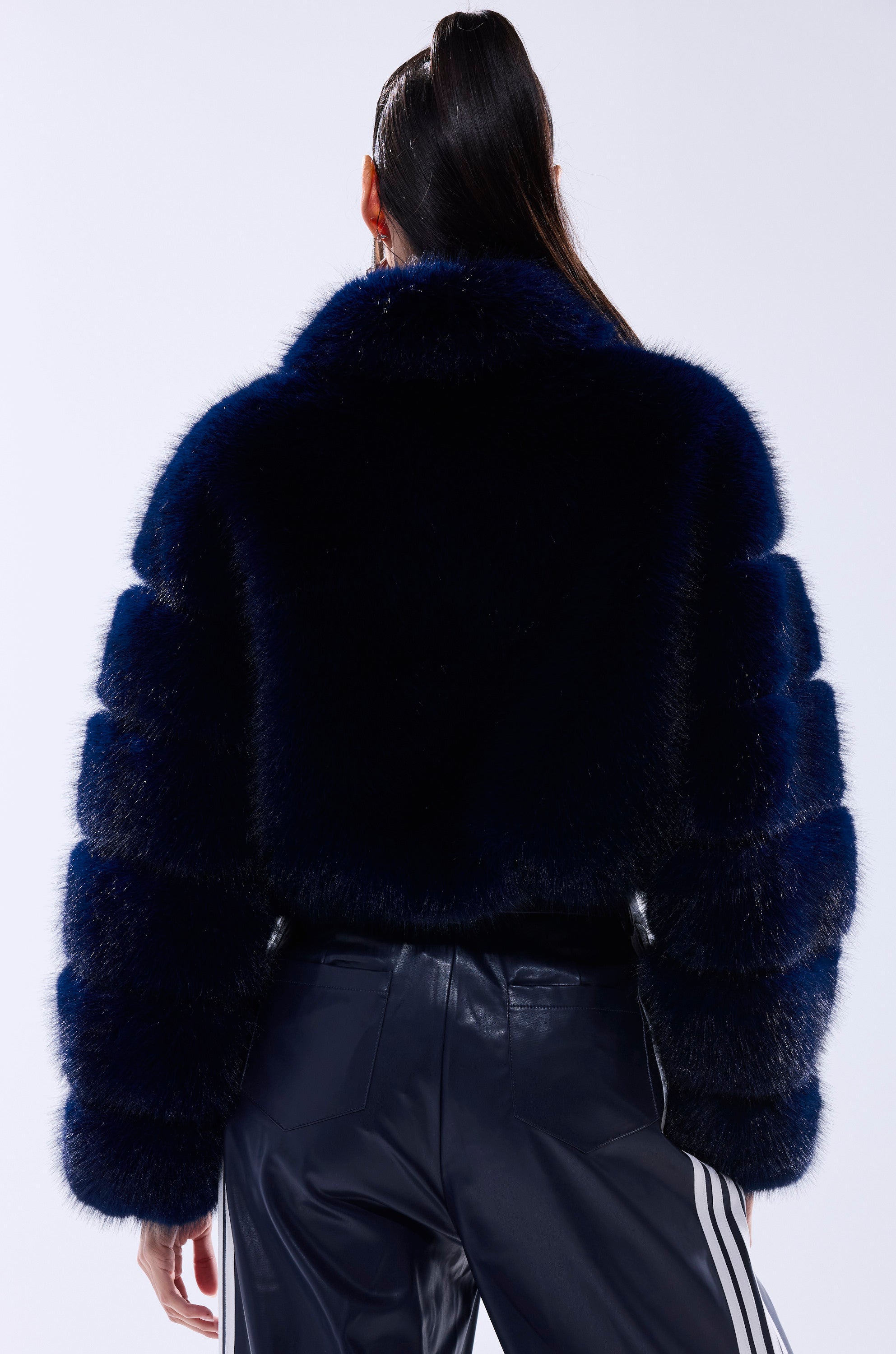 A person with long dark hair in a high ponytail is seen from behind wearing a cropped KEIKO FAUX FUR COAT in dark blue and shiny pants with white side stripes, set against a plain light-colored background.