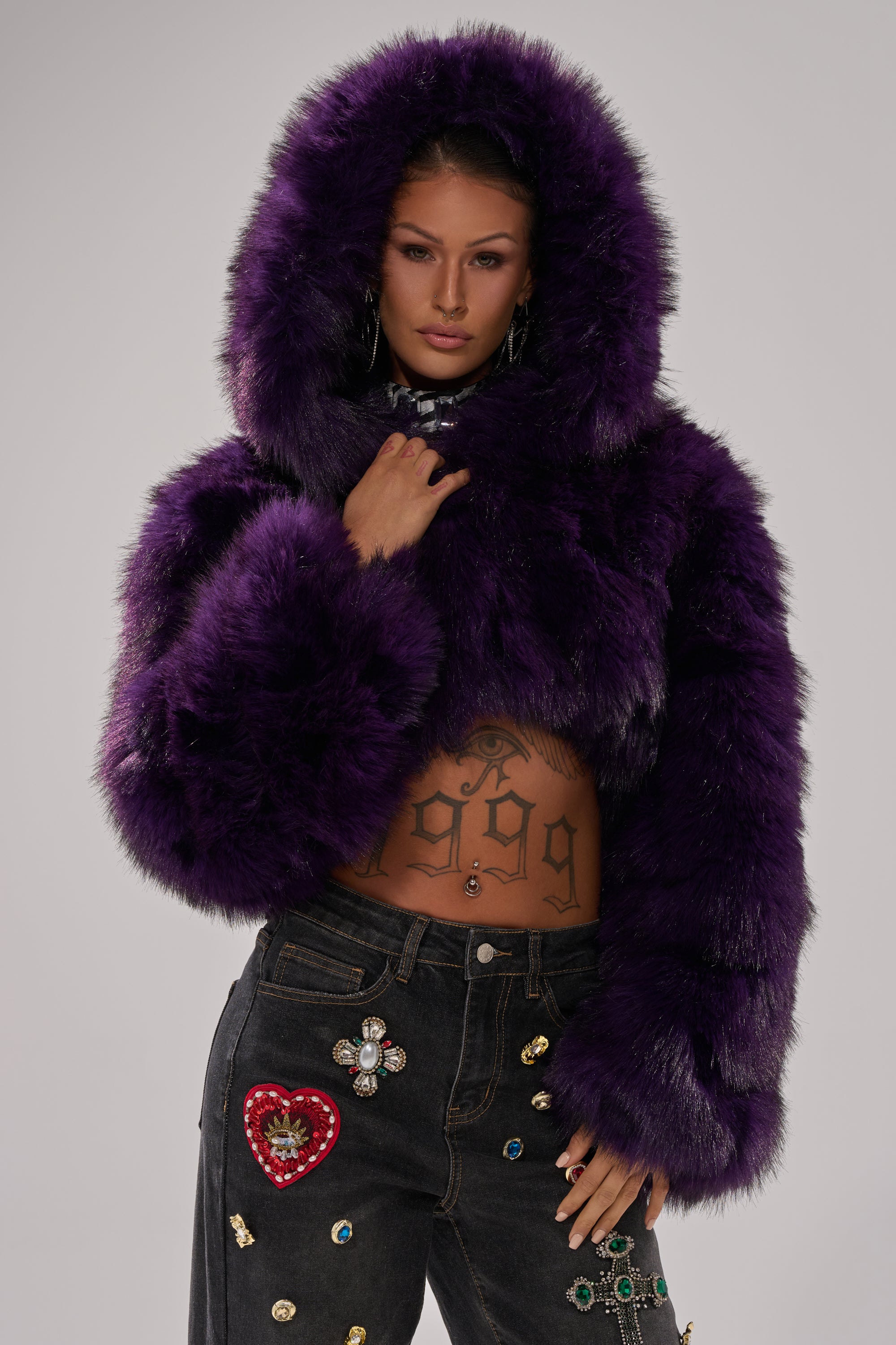 Wearing the JUST A FLING CROPPED FAUX FUR jacket in deep purple and black jeans with colorful patches and rhinestones, a person poses confidently, showing off a "1999" tattooed midriff.