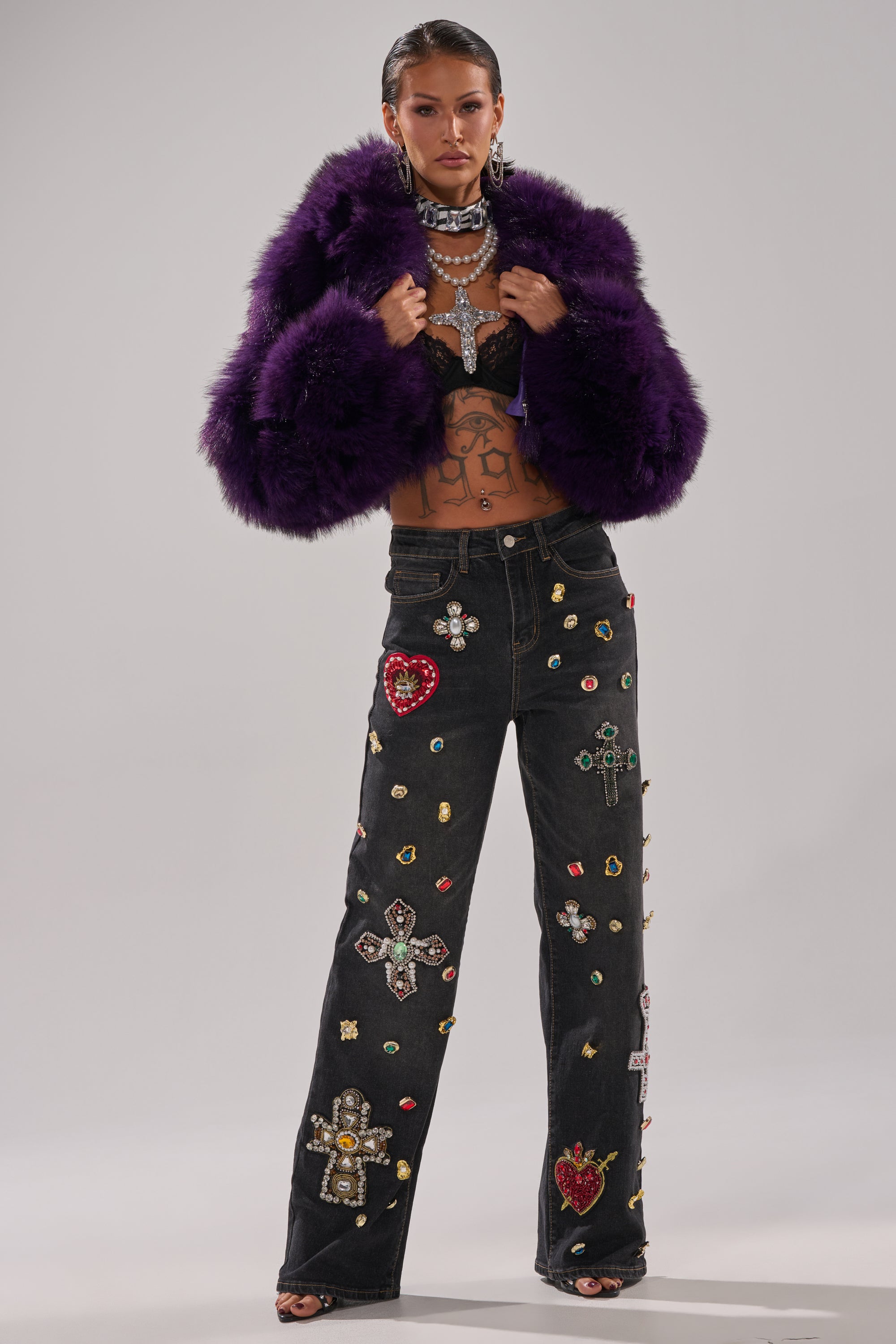 A woman exudes confidence in the JUST A FLING CROPPED FAUX FUR jacket, a black bralette, statement jewelry, and black jeans featuring colorful jeweled patches shaped like crosses and hearts.