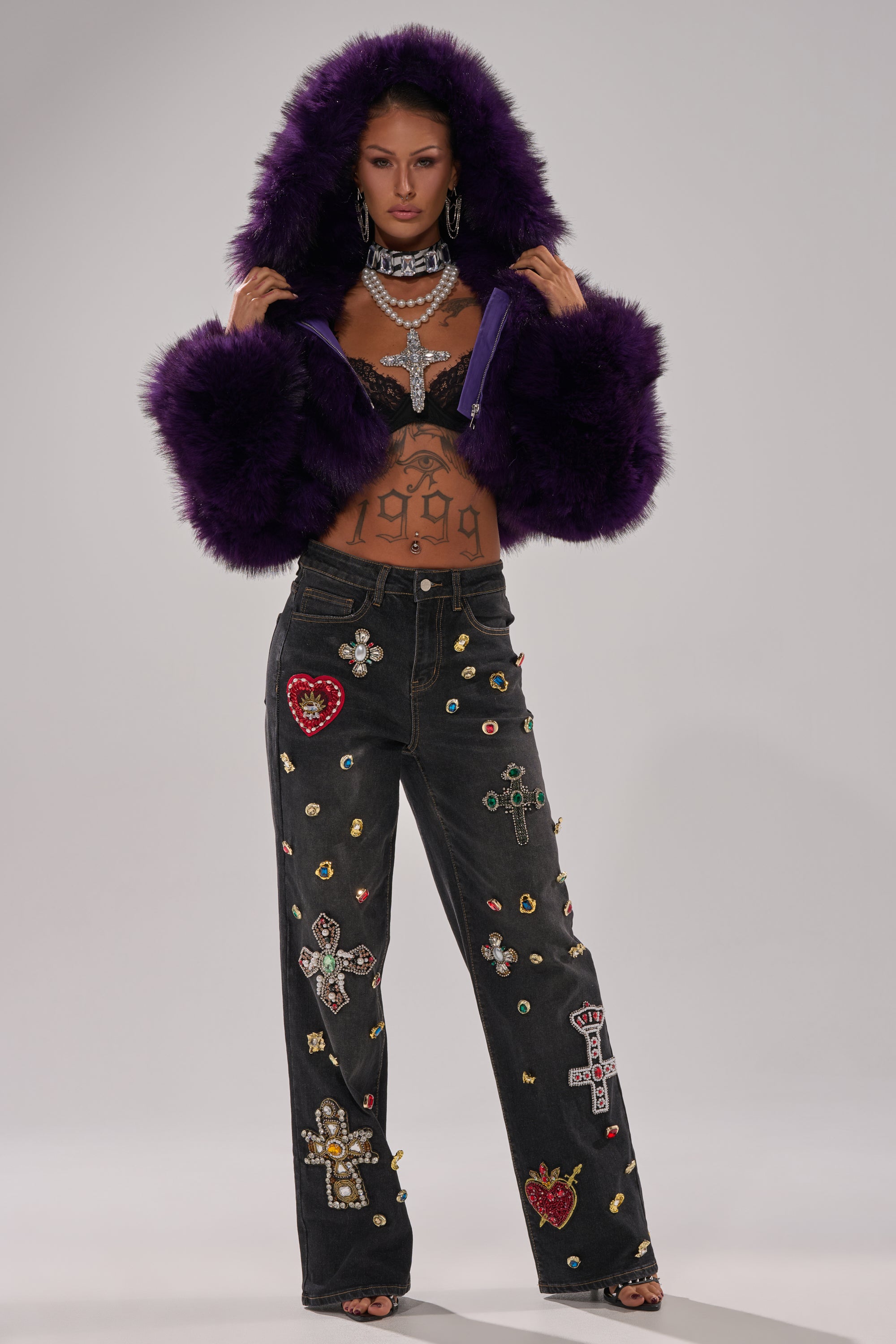 A woman stands facing forward, wearing the JUST A FLING CROPPED FAUX FUR jacket with a hood, jeweled bralette, statement necklace, and high-waisted black jeans decorated with colorful gems and cross patches.
