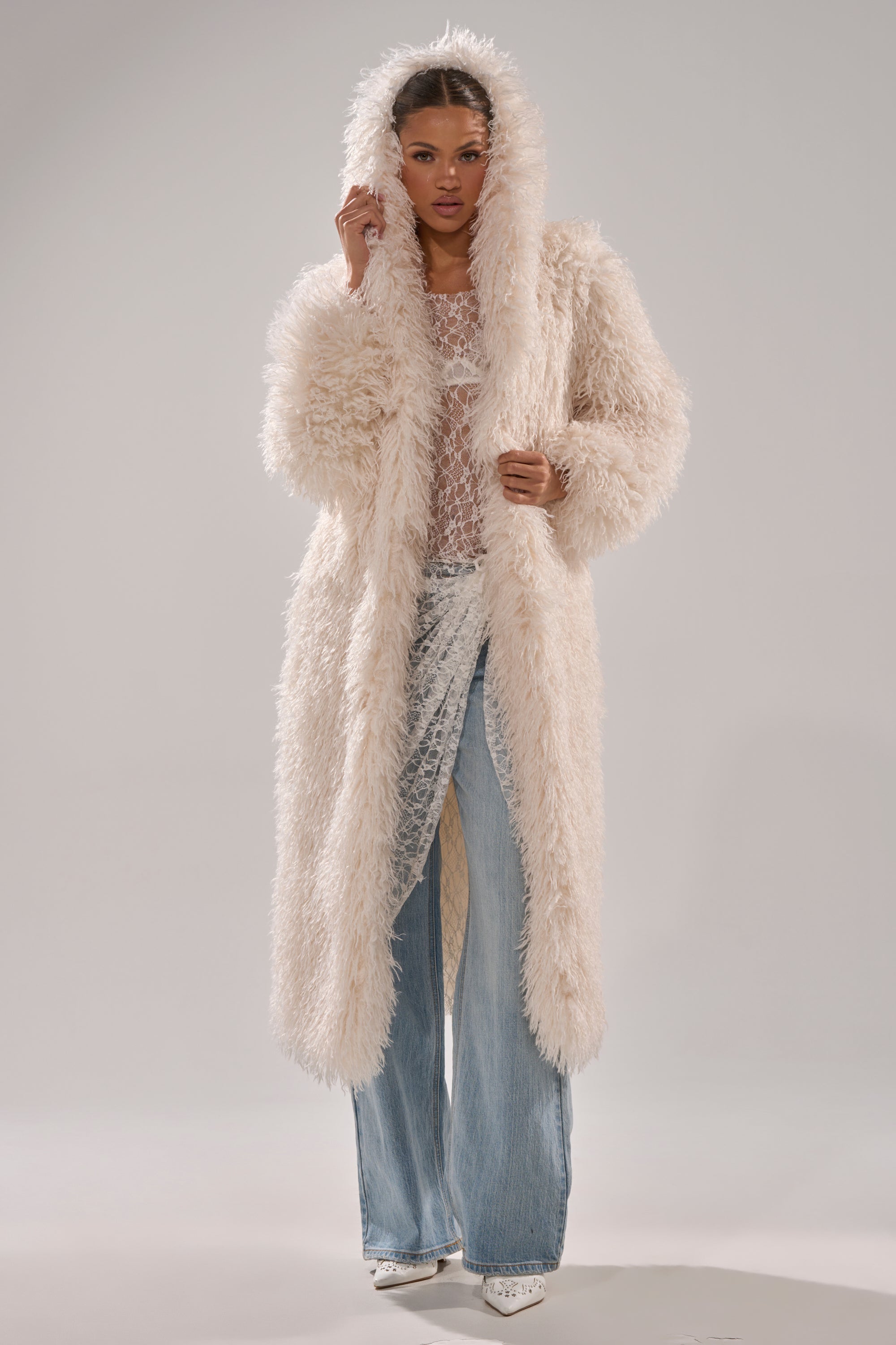 A woman wears the SCARLETT FAUX FUR COAT with a hood over a sheer top and wide-leg blue jeans, standing against a plain light background.