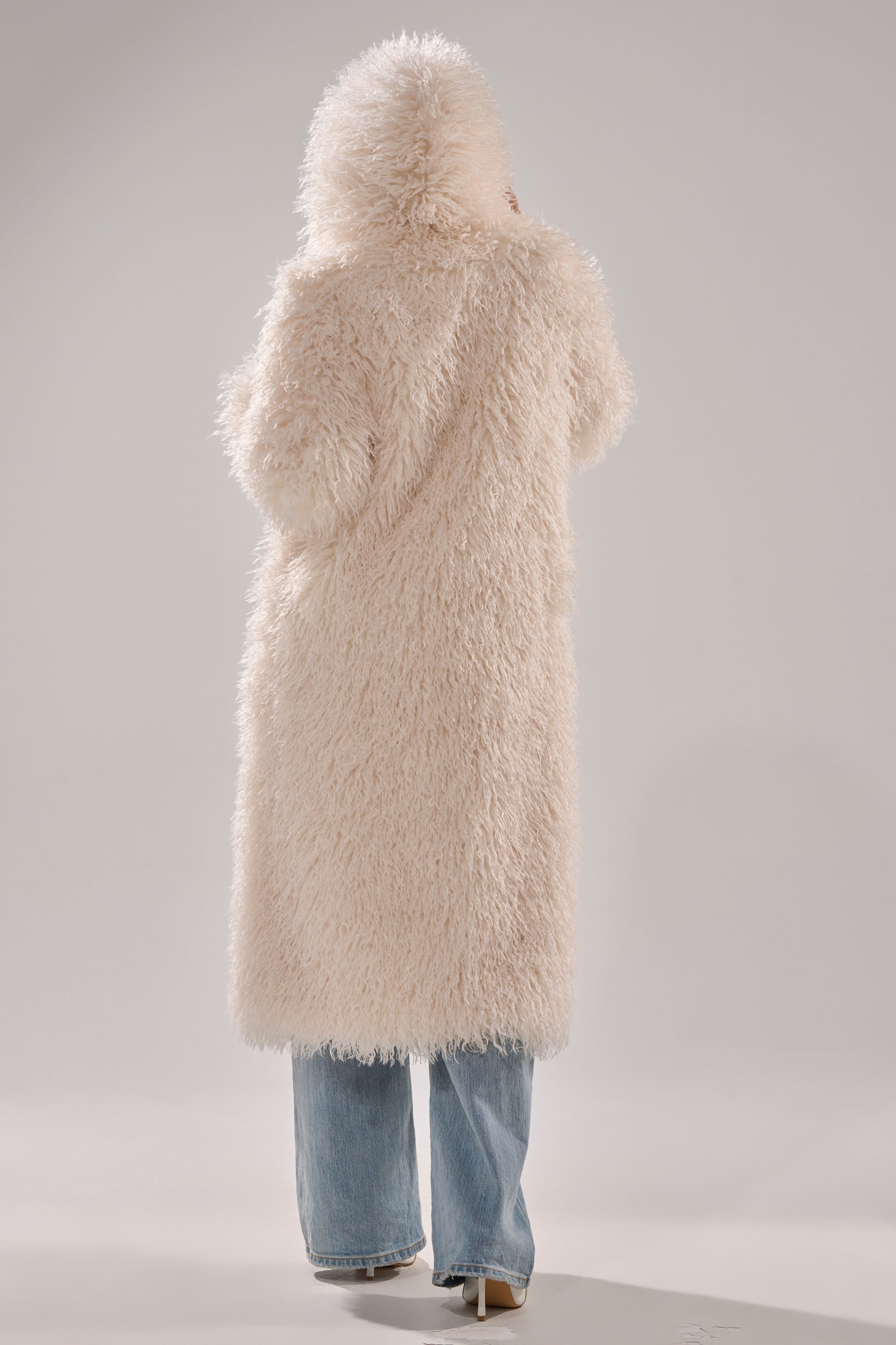 A person in the SCARLETT FAUX FUR COAT, featuring a shaggy-chic cream design and oversized silhouette, pairs it with blue jeans while facing away from the camera against a plain background.