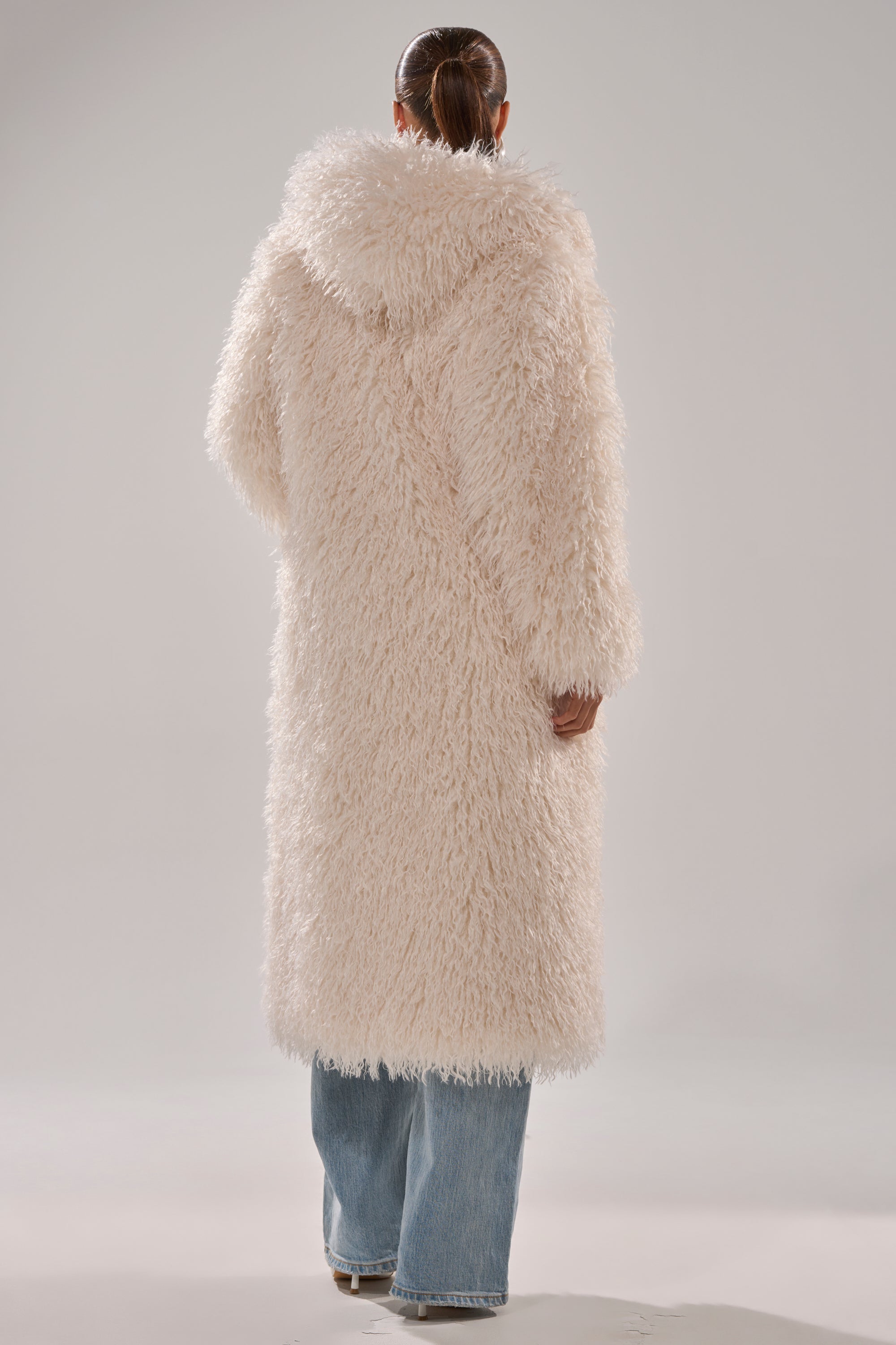 A person stands facing away, modeling the SCARLETT FAUX FUR COAT—an off-white, hooded, oversized silhouette with a shaggy-chic look—paired with wide-leg blue jeans and white shoes. Their hair is styled in a low ponytail.