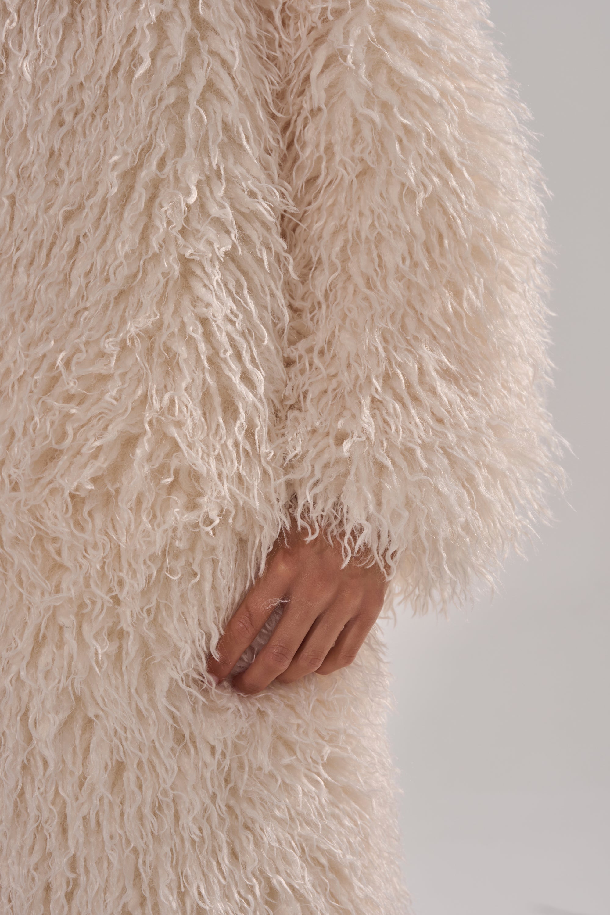 A close-up of a hand resting on the SCARLETT FAUX FUR COAT, featuring an oversized fit and fluffy off-white shaggy texture, set against a plain light background.