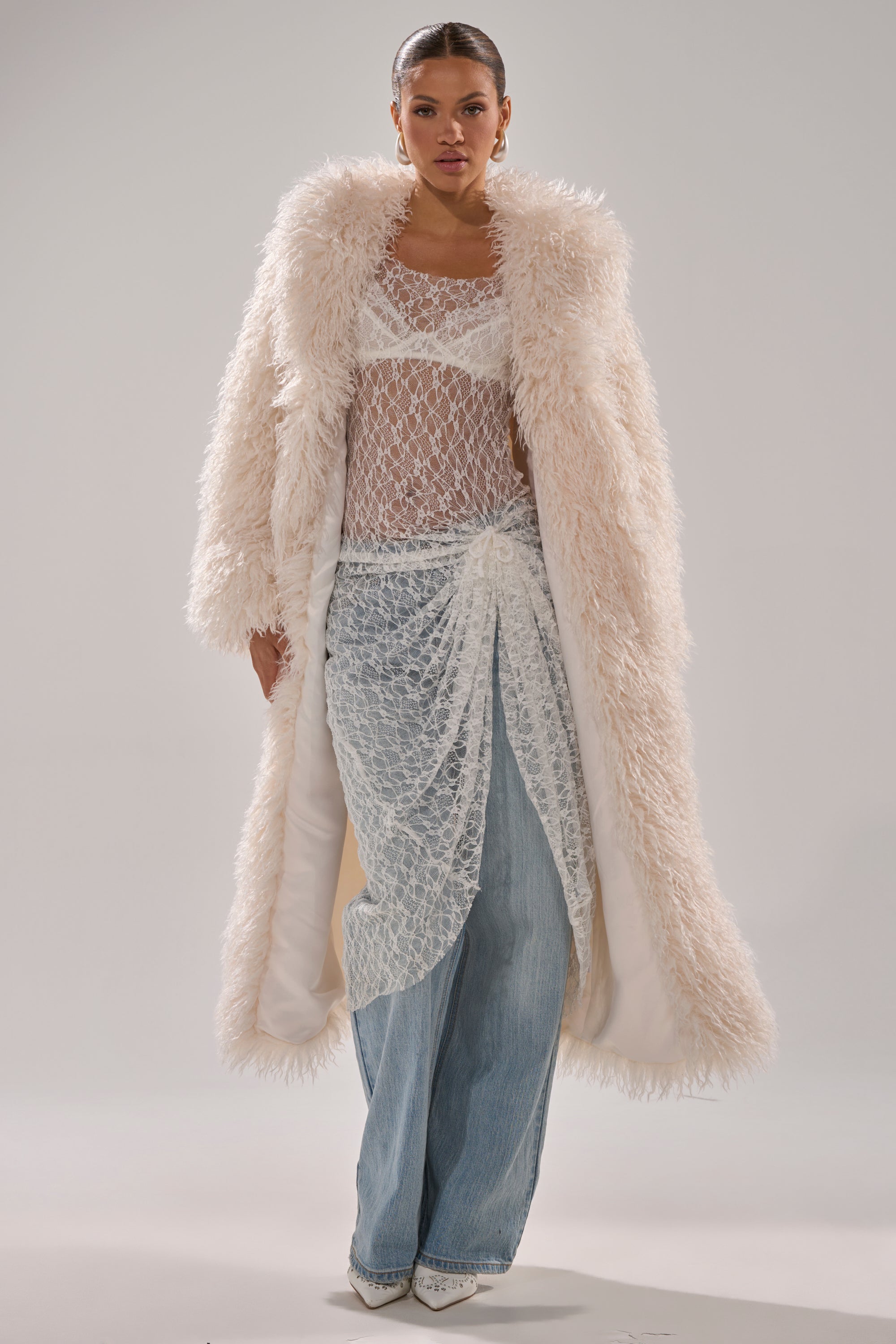 A woman models the SCARLETT FAUX FUR COAT over a sheer white lace top and skirt, styled with light blue wide-leg jeans and pointed-toe shoes, against a plain background.