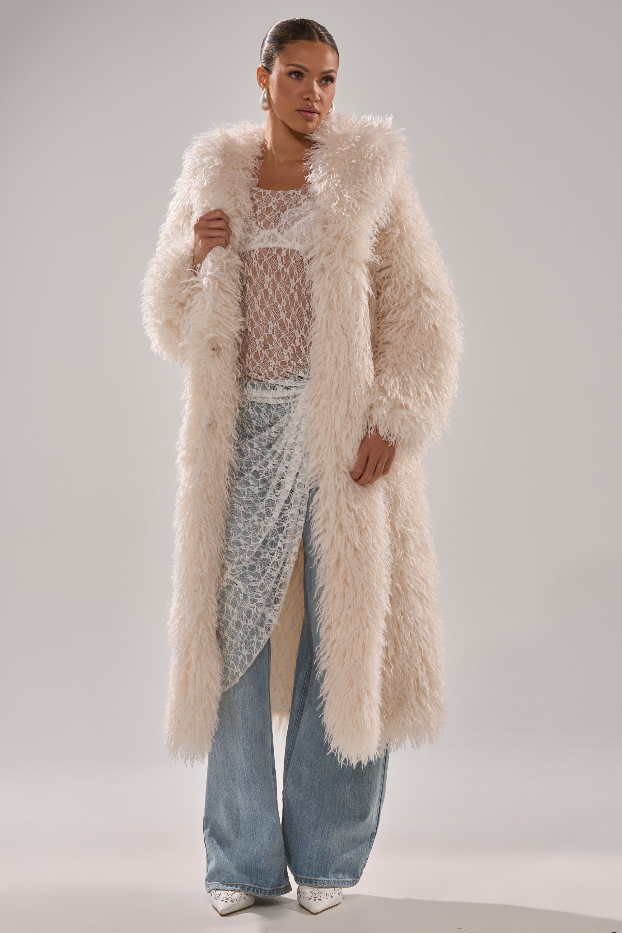 A woman stands against a plain backdrop wearing the SCARLETT FAUX FUR COAT over a sheer textured top and light blue wide-leg jeans. With her hair pulled back, she poses confidently, looking slightly to the side.