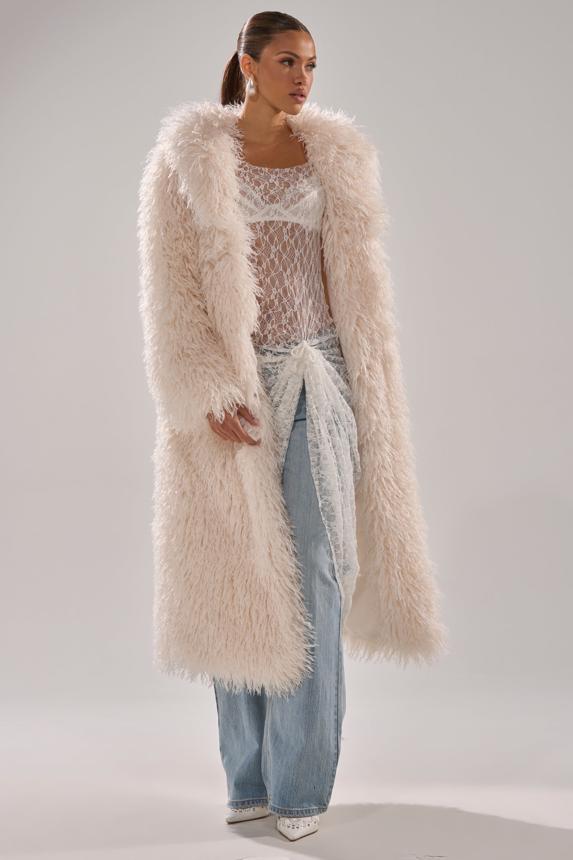 A woman wears the SCARLETT FAUX FUR COAT over a sheer white top, light blue wide-leg jeans, and white pointed-toe shoes against a plain light background.