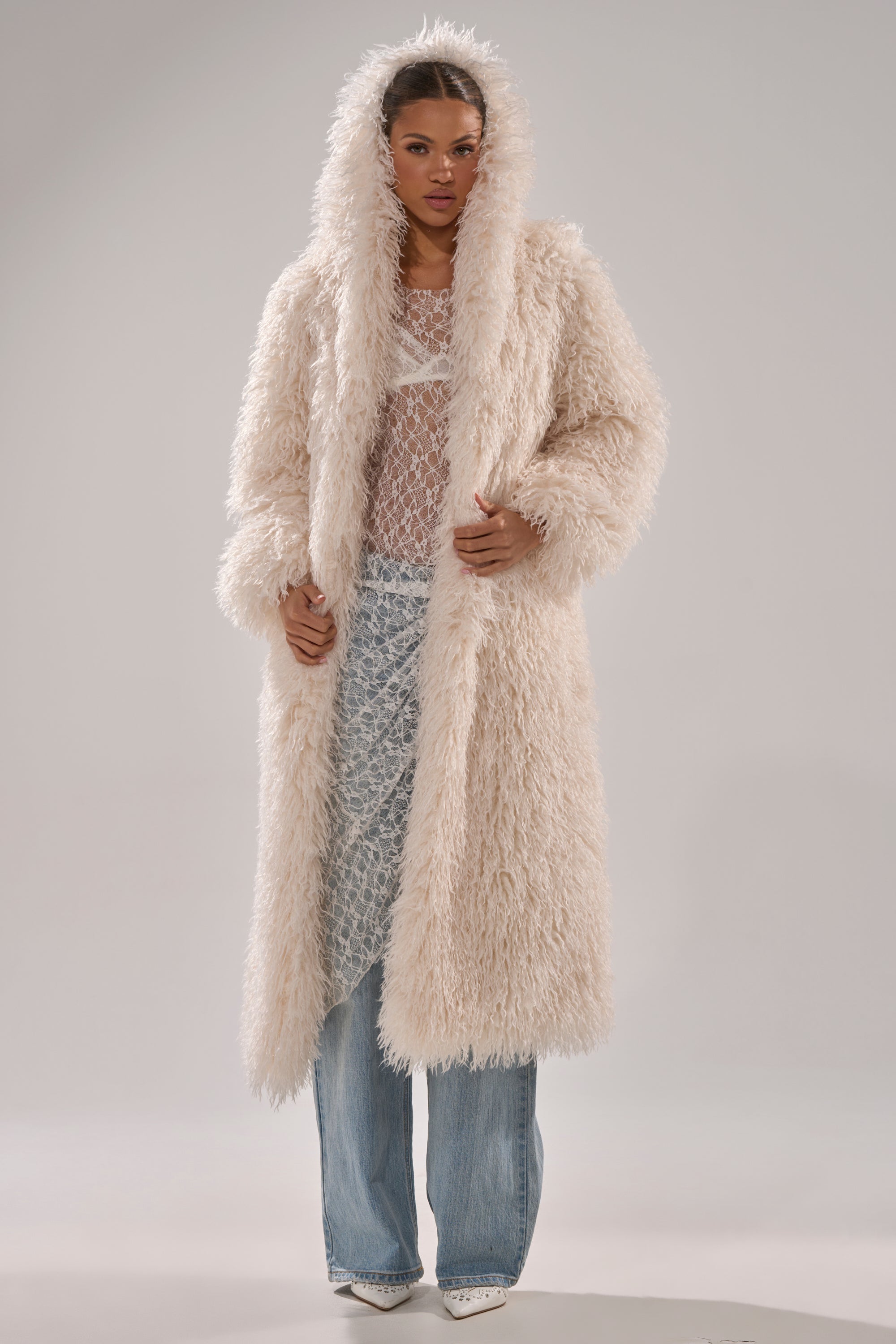 A woman stands against a plain light background wearing the SCARLETT FAUX FUR COAT—a long, oversized faux fur coat with a hood—over a sheer lace top, blue jeans, and pointed white shoes.