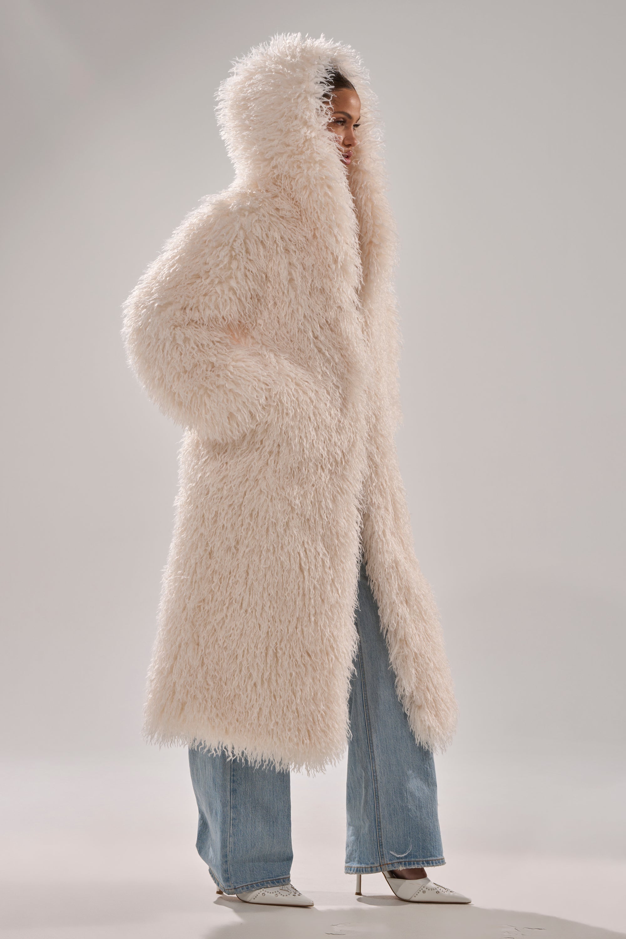 A person wearing the SCARLETT FAUX FUR COAT with a hood, blue jeans, and white pointed-toe heeled boots stands against a plain light gray background.
