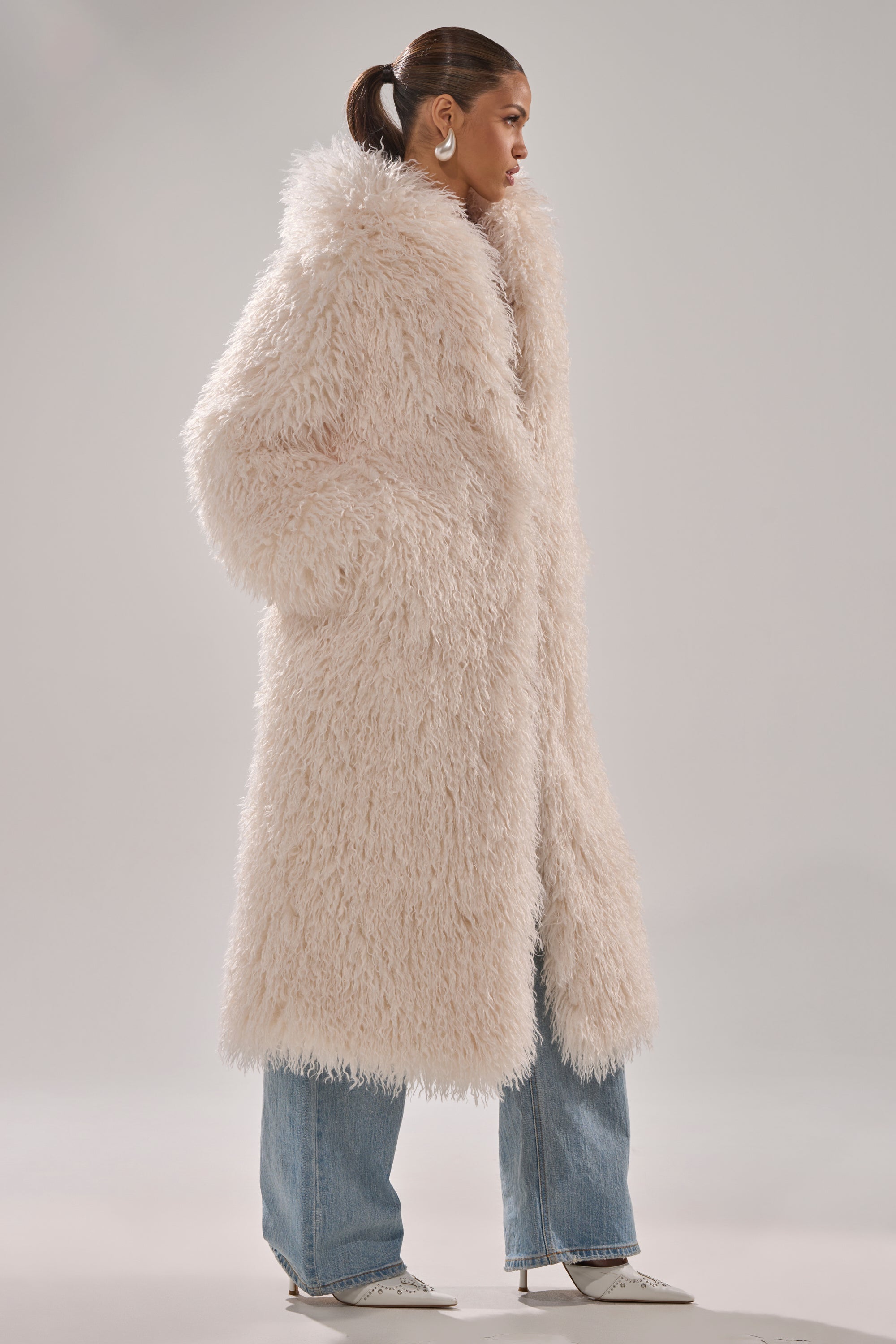 A woman stands in profile wearing the SCARLETT FAUX FUR COAT, light blue jeans, pointed white heels, and hoop earrings against a simple gray background.