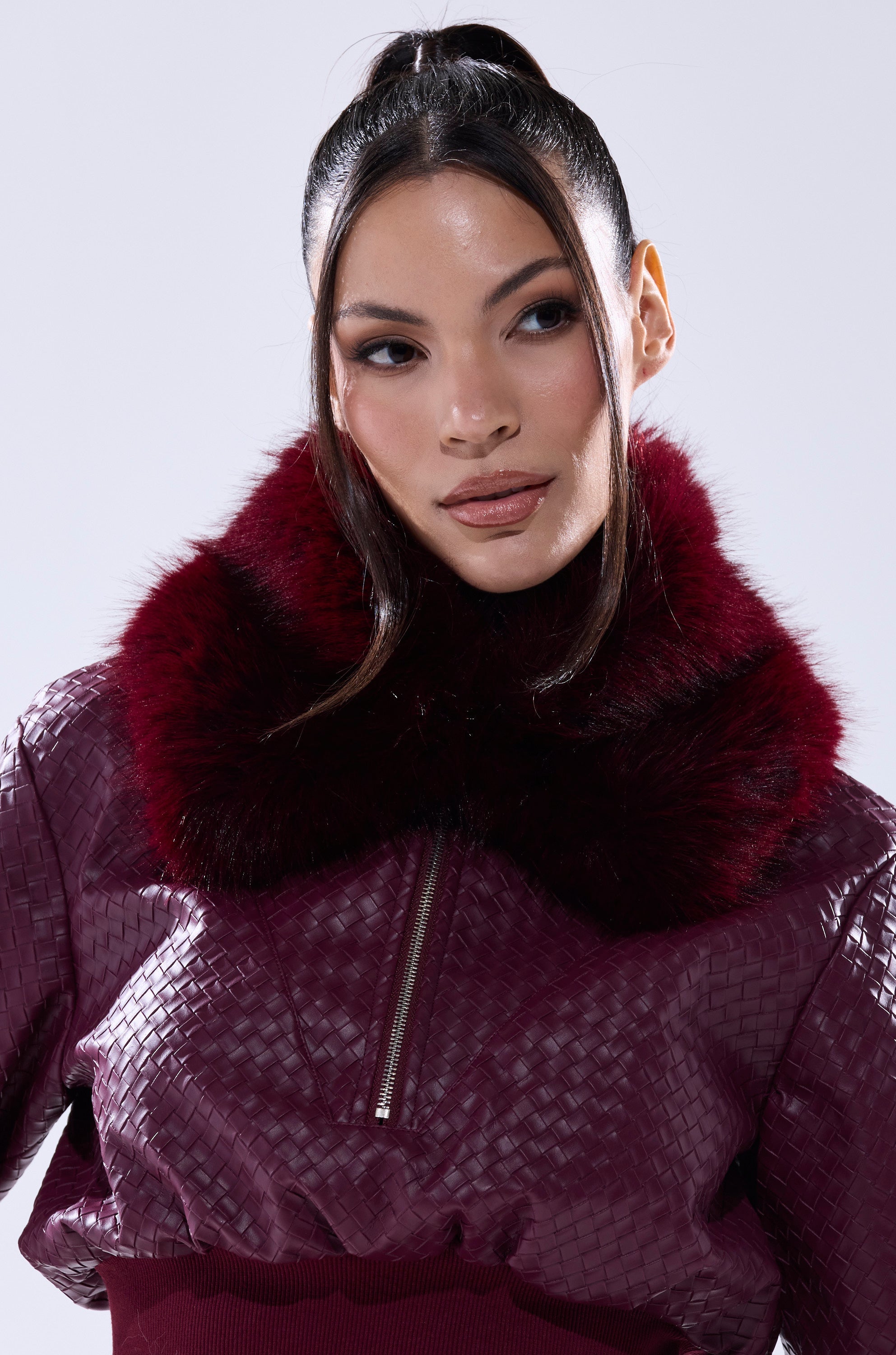 A woman with long dark hair in a high ponytail wears the ALAIA FAUX FUR COLLAR, a textured jacket with a large, fluffy burgundy faux fur collar. She looks slightly to the side against a plain light background. 0
