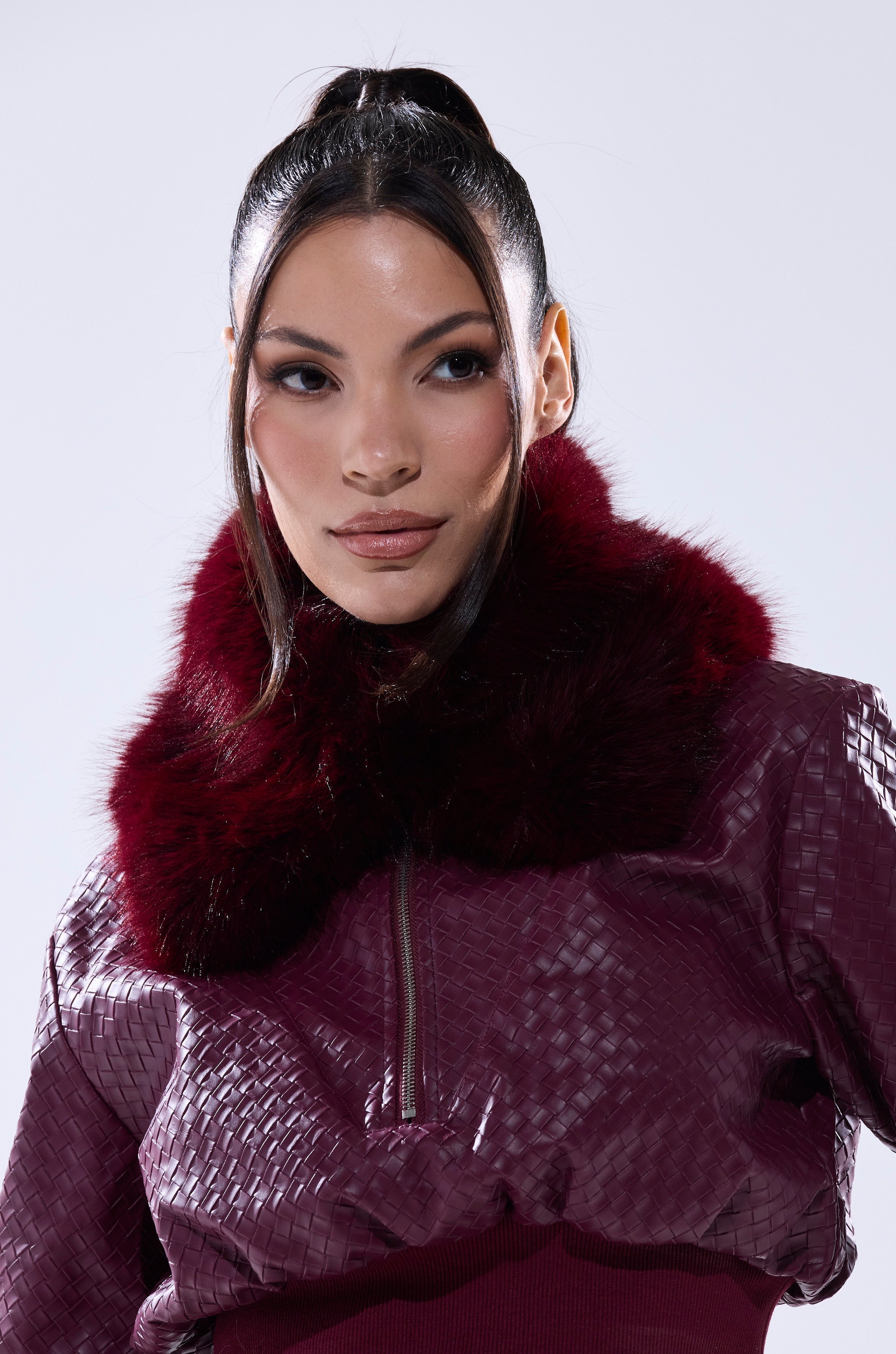 A woman with long dark hair in a high ponytail wears the ALAIA FAUX FUR COLLAR jacket, which features a shiny burgundy textured finish and a thick faux fur collar, posing against a light gray background. 1