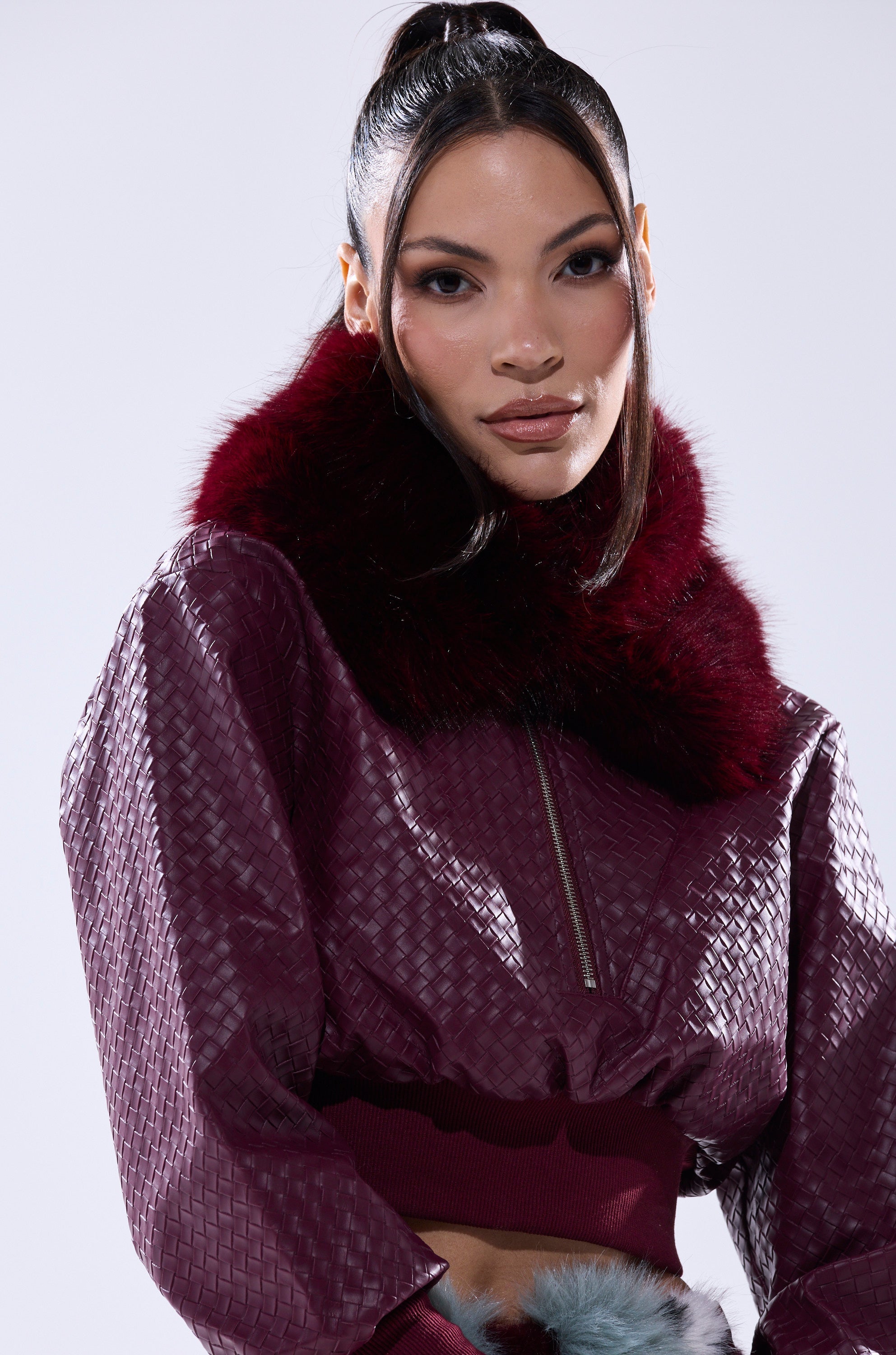 A woman with long dark hair in a high ponytail confidently poses against a plain light background wearing the AKIRA Label ALAIA FAUX FUR COLLAR textured burgundy jacket with a large dark red faux fur collar. 3