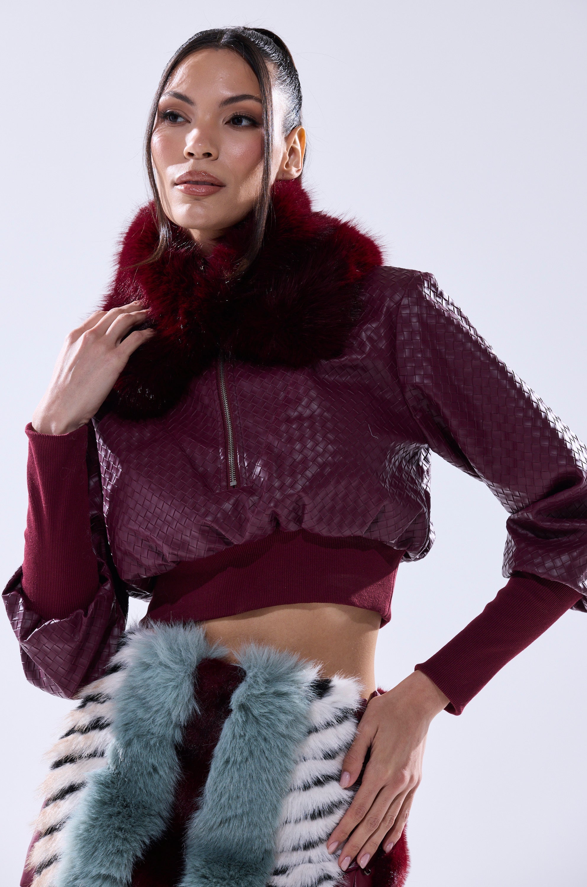 A woman poses in an ALAIA FAUX FUR COLLAR crop jacket with a textured, shiny finish and deep burgundy faux fur collar, styled with a skirt adorned with fluffy multicolored blue, white, black, and red fur panels. 4
