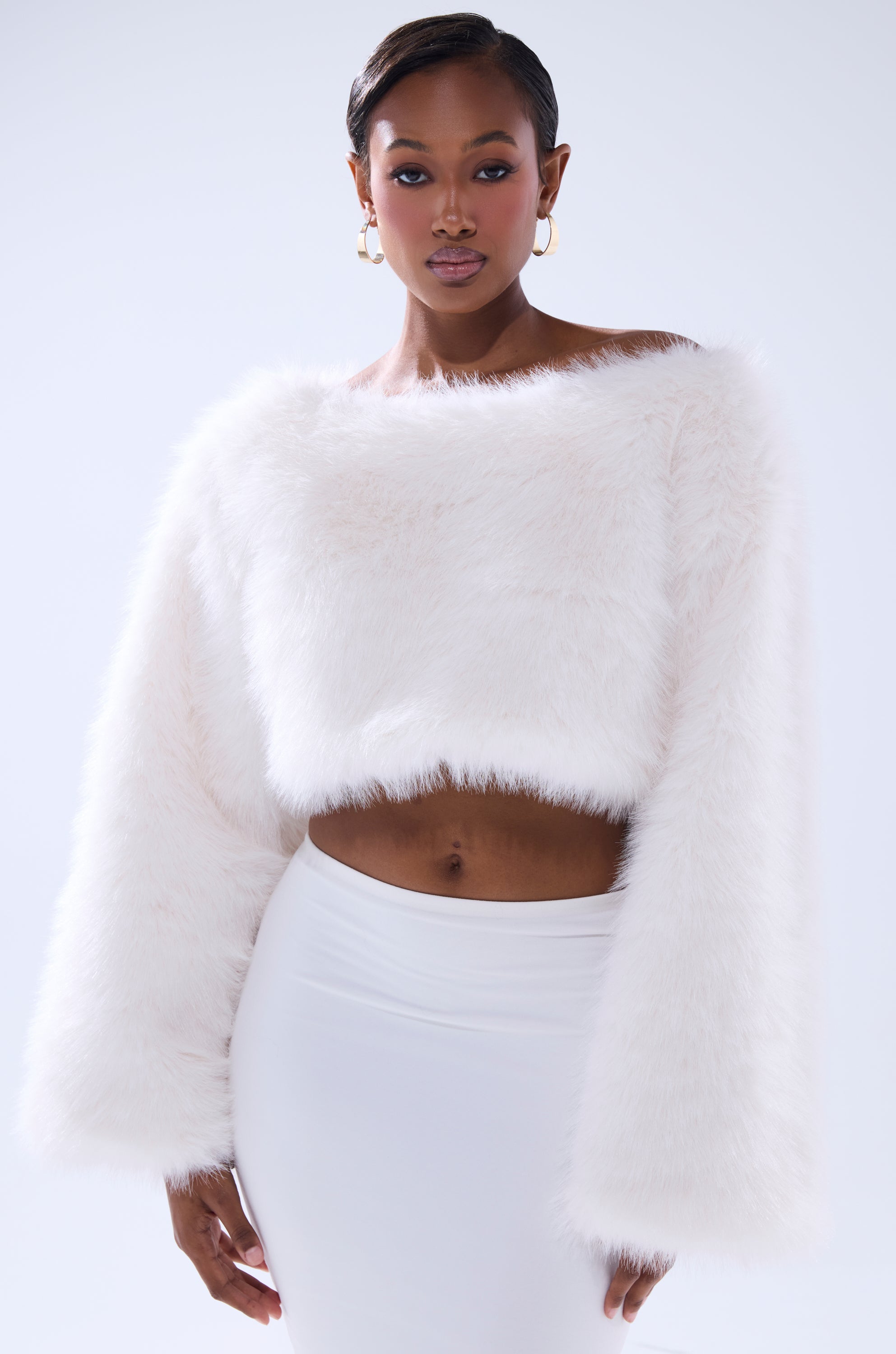 A woman with short hair and hoop earrings confidently poses against a plain light background wearing the COMFY COZY CROPPED SWEATER, a fuzzy white faux fur crop top, paired with a fitted white skirt.