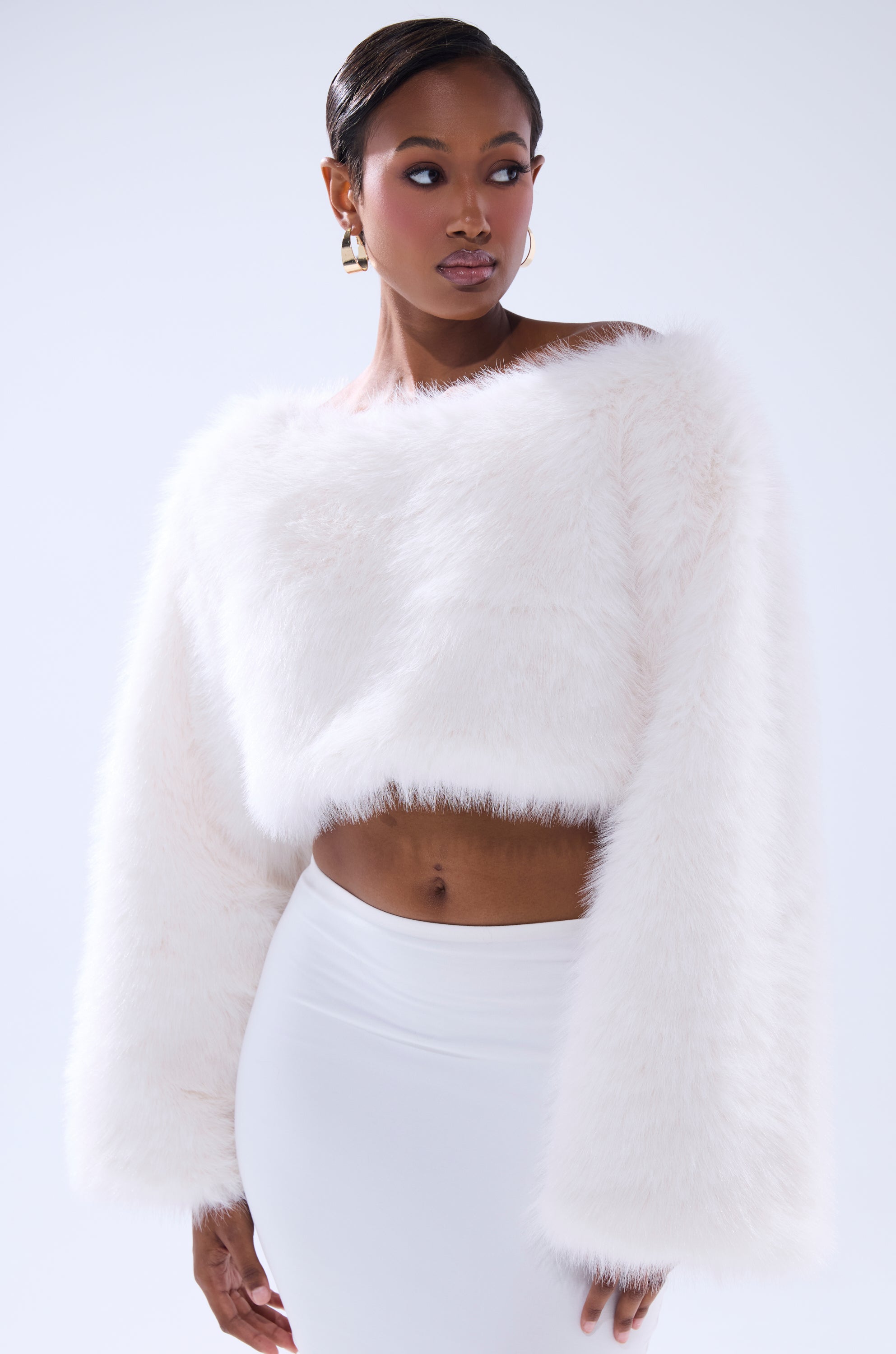 A woman wears the COMFY COZY CROPPED SWEATER, a white faux fur long-sleeve piece, paired with a fitted white skirt as she stands against a light background, glancing to the side with a neutral expression.