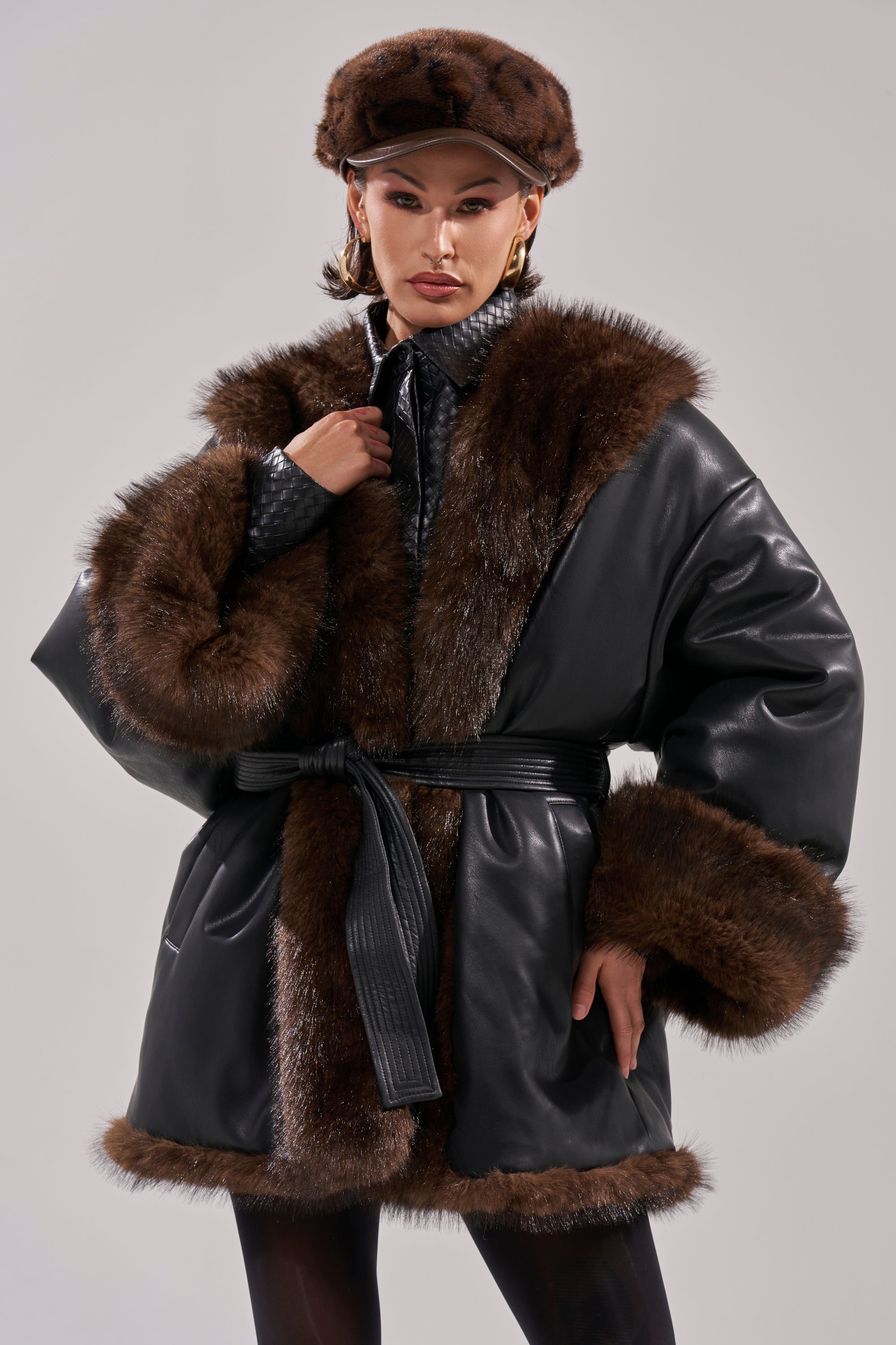 A person poses confidently in the CAN I KICK IT REVERSIBLE PUFFER, featuring a luxurious faux leather finish with wide brown fur trim, paired with a matching fur cap and statement earrings against a plain light background.
