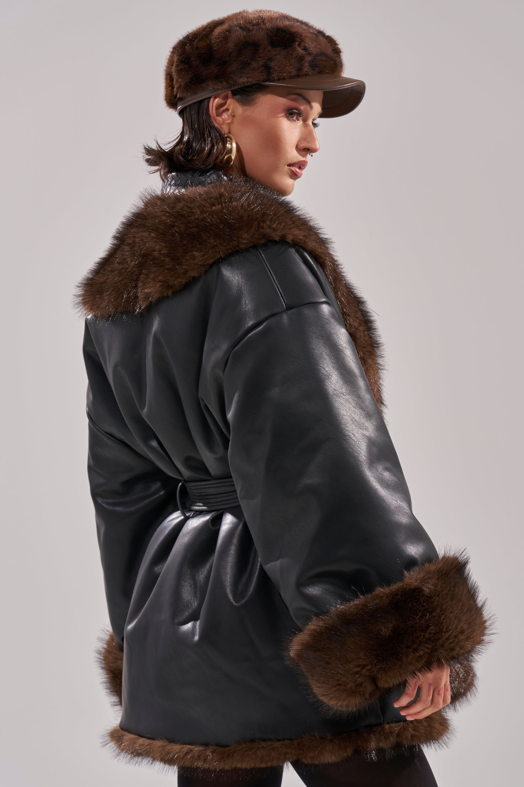 Wearing the CAN I KICK IT REVERSIBLE PUFFER, a black faux leather coat with brown fur trim, and a matching brown fur hat, a person poses against a plain light background, looking over their shoulder in chic style.