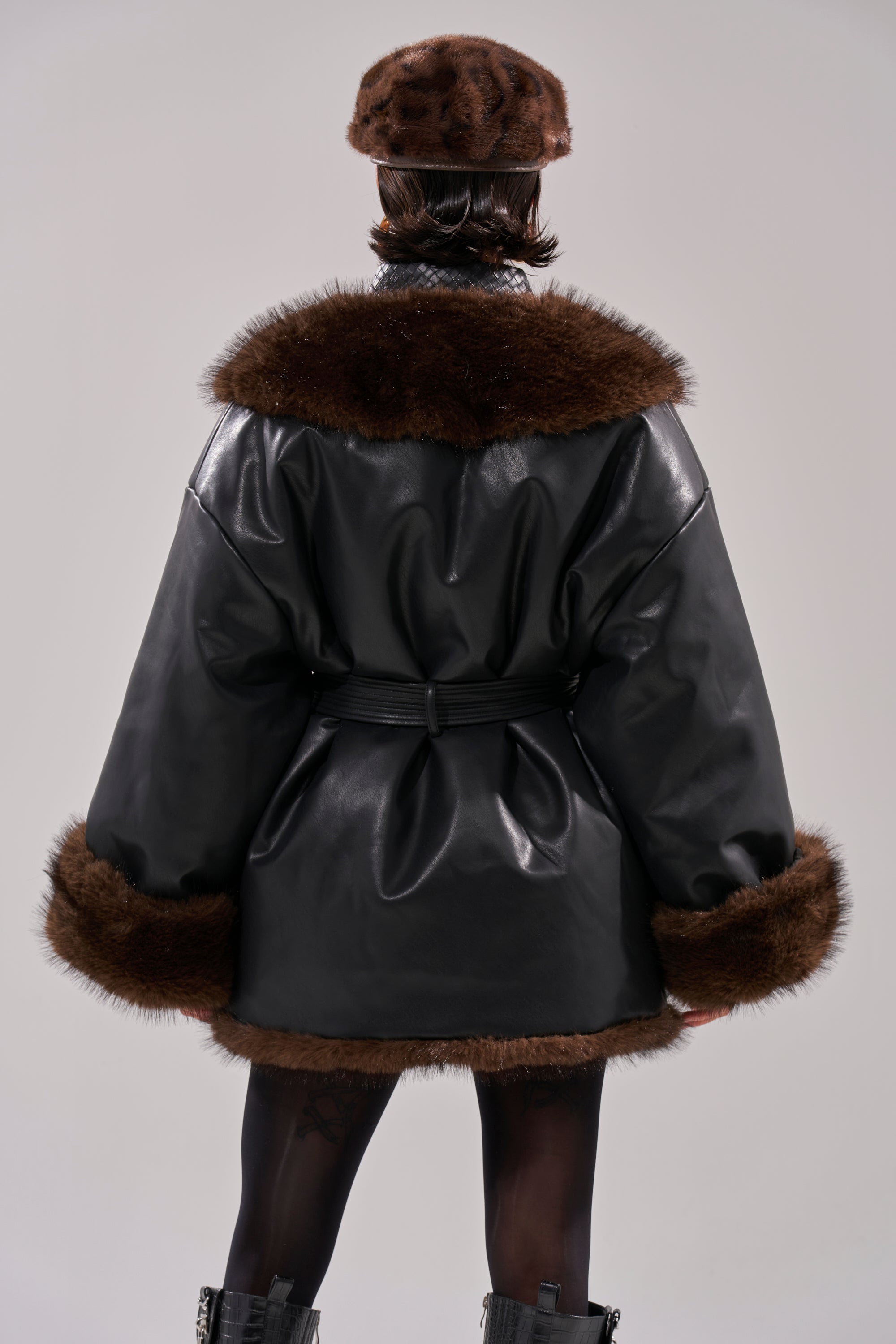 A person stands facing away, wearing the CAN I KICK IT REVERSIBLE PUFFER with dark brown fur trim, a matching beret, black tights, and black boots against a plain light background.