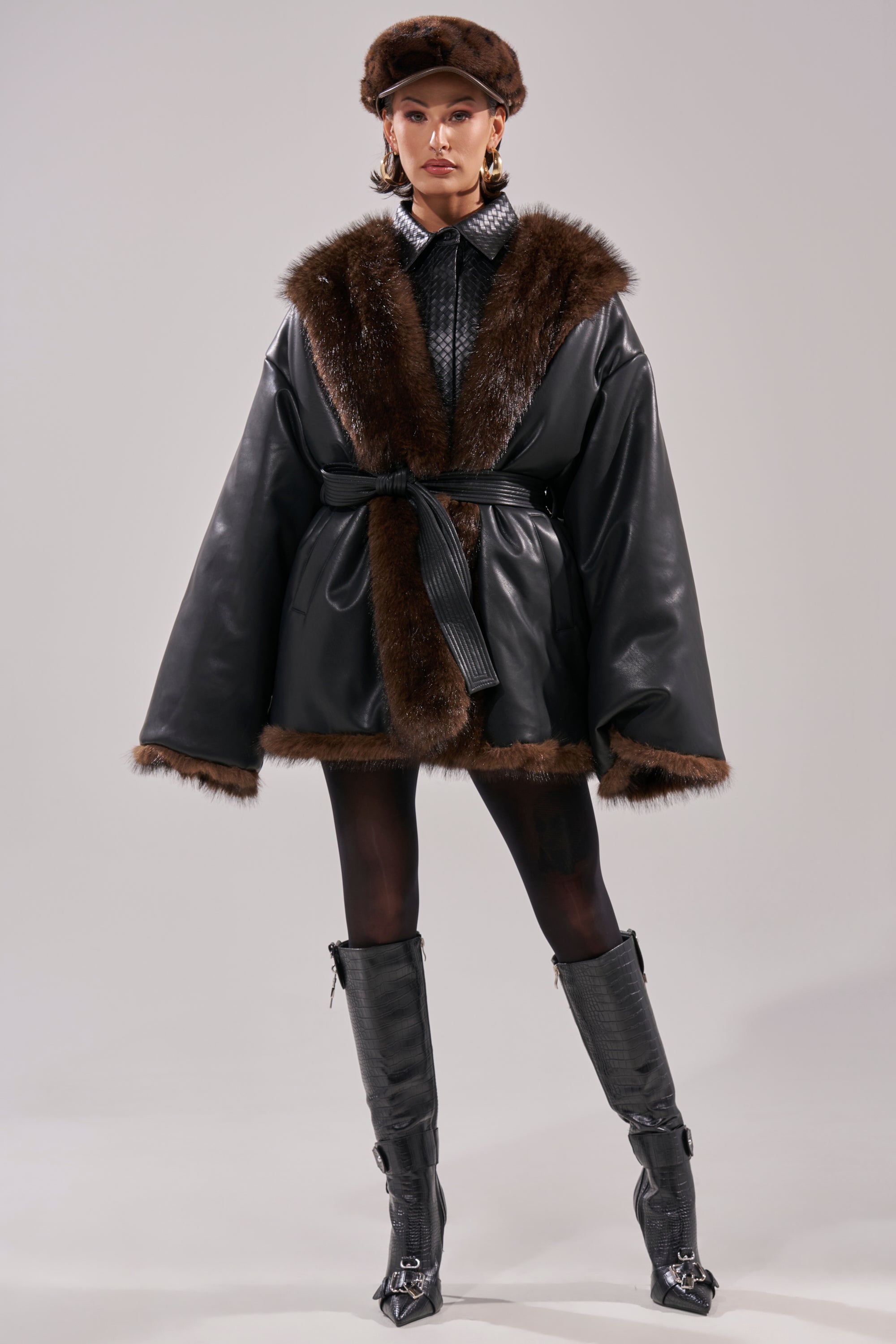 A woman faces forward, wearing the CAN I KICK IT REVERSIBLE PUFFER—a wide black faux leather coat with brown faux fur trim—paired with a matching fur hat, black tights, tall boots, gold hoop earrings, and a confident look.