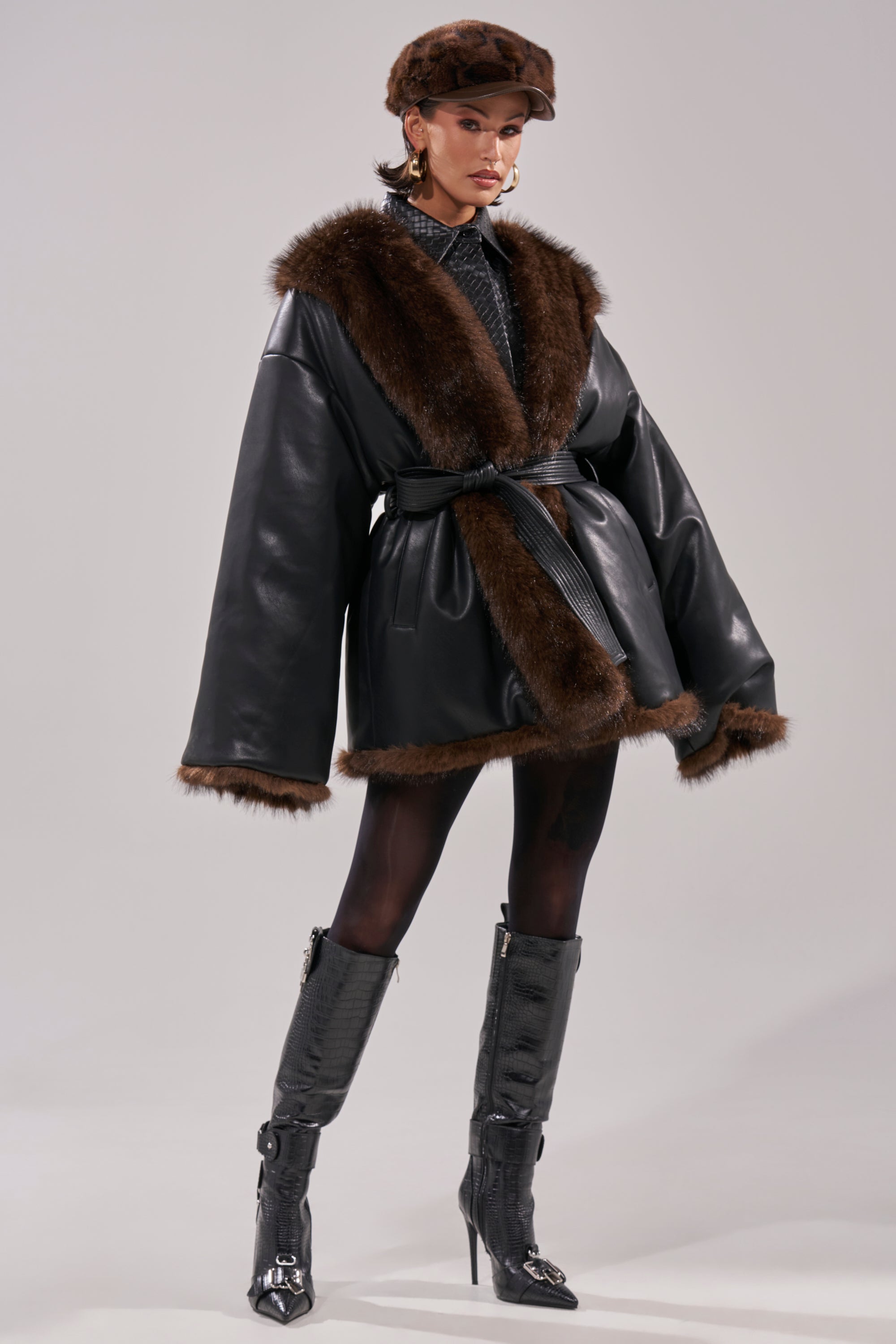 A person models the CAN I KICK IT REVERSIBLE PUFFER, styled with a brown fur hat, tall textured boots, and dark tights against a plain background.